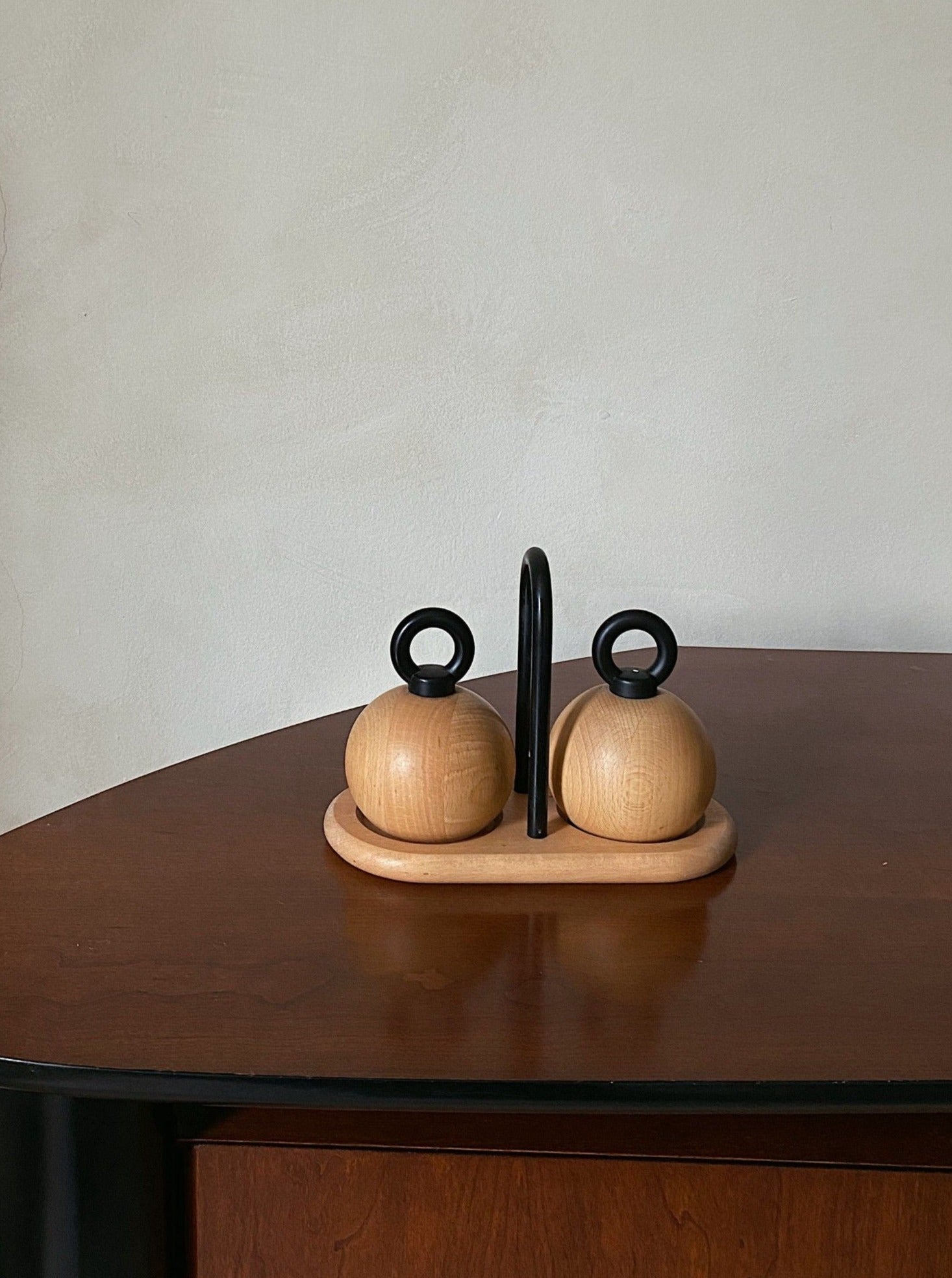 A minimalist Bodum C. Jörgensen Salt & Pepper set, designed by Les Objets Amsterdam, consisting of two round wooden shakers with black circular handles, symmetrically placed on a wooden table against a plain beige wall