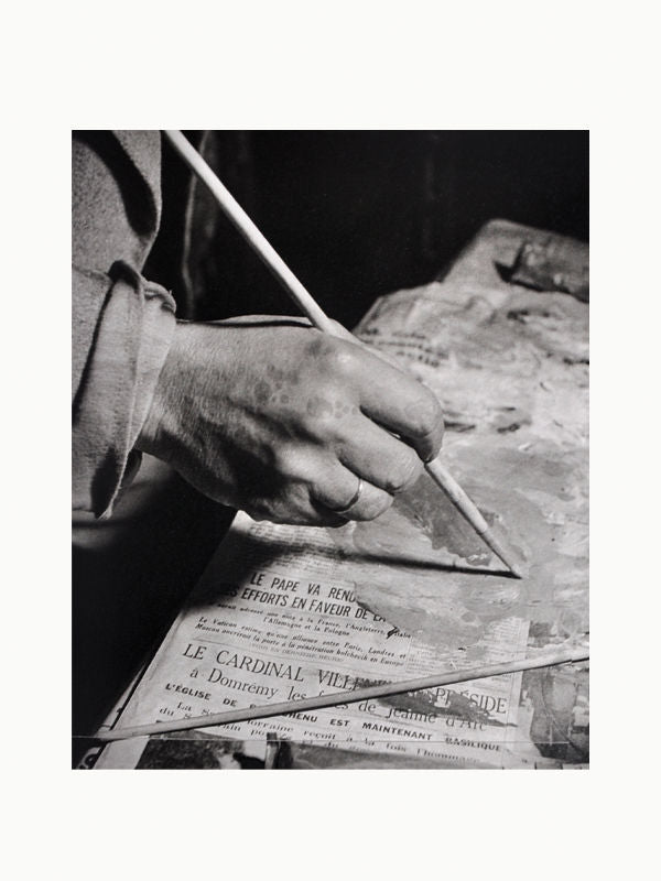 Close-up black and white image of a person's hands holding a pencil and writing on a newspaper. The visible text on the newspaper is in French, capturing the essence of the Paris art scene with Maison Plage's Brassai: Paris & Picasso.