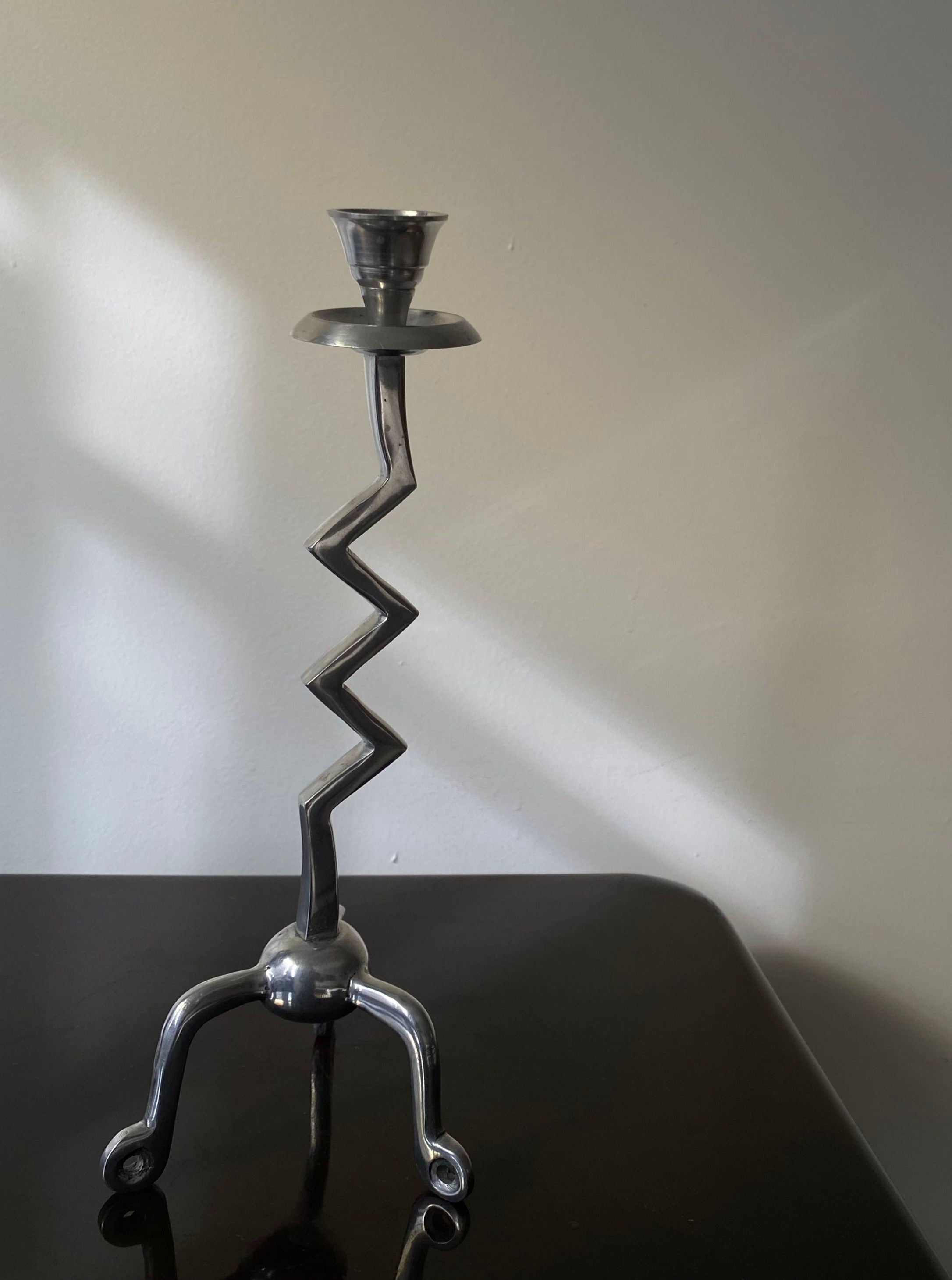 The Vintage Zigzag Candle Holder by Les Objets Amsterdam is a handcrafted metallic piece with a zigzag stem, three-leg base, and candle cup. Its contemporary design enhances luxury interiors, standing beautifully on dark surfaces against light-colored walls.