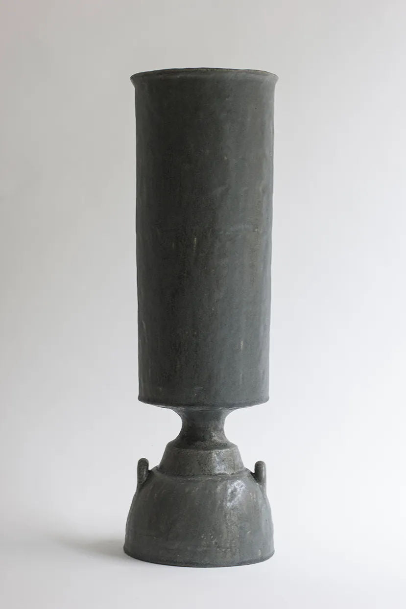 The Isolated n.32 Vase by Canoa Lab is a tall, textured dark gray piece with a cylindrical upper section and rounded base on a smooth white background. It exemplifies contemporary design through its minimalist aesthetic, featuring subtle handles on the base.