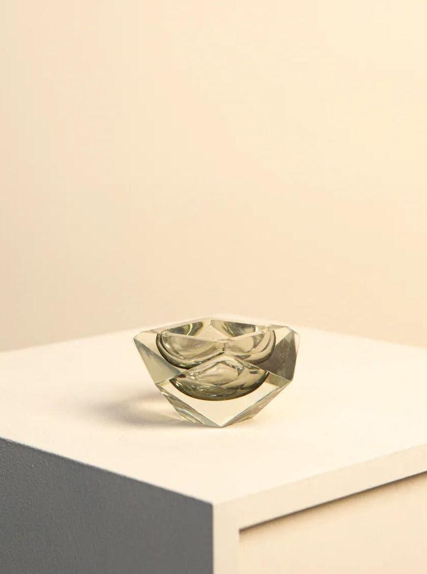 A small, transparent Treaptyque ashtray rests symmetrically on a white surface against a soft beige background. The ashtray, designed by Flavio Poli for Seguso, has