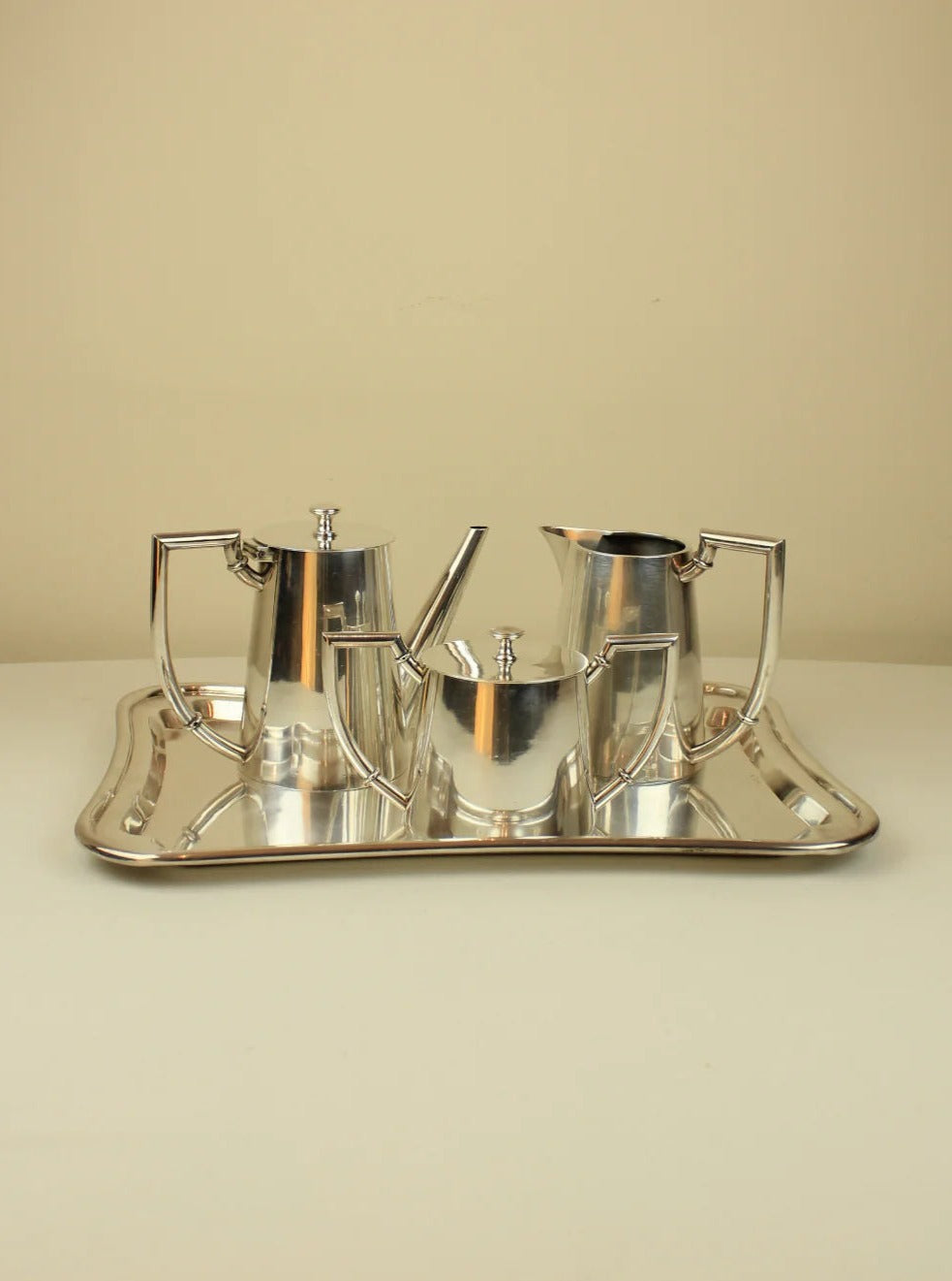 A set of modern stainless steel Art Deco Tea/Coffee Service on a tray, including a teapot, creamer, sugar bowl, and a serving pot, displayed against a plain beige background by Boga Avante Shop.