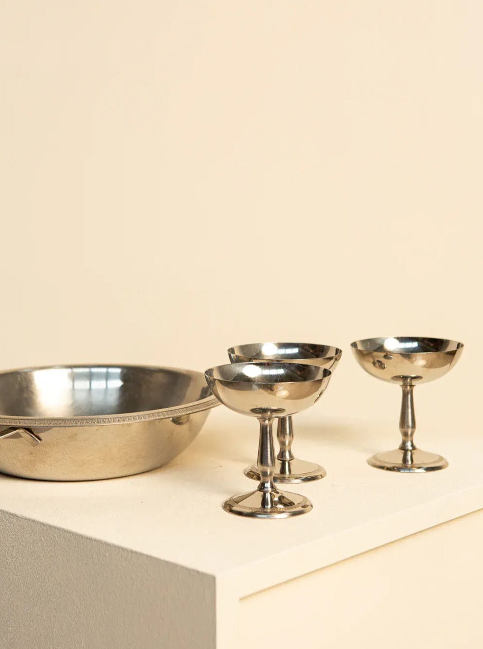 Four silver bowls from Treaptyque's Set of 6 Ice Cream Bowls and a 60's Basket are artfully arranged on a cream-colored pedestal against a beige background. One bowl is larger and flatter, while the other three are smaller with rounded bodies and short stems, embodying a retro design that exudes vintage charm. They resemble elegant Italian ice cream bowls from the 1960s.