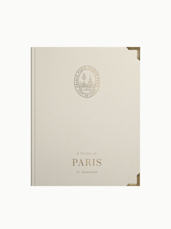 A beige hardcover book titled "Paris Travel Guide By Seasons," featuring a simplistic design and an emblem with a tree and buildings, on a white background by Maison Plage.