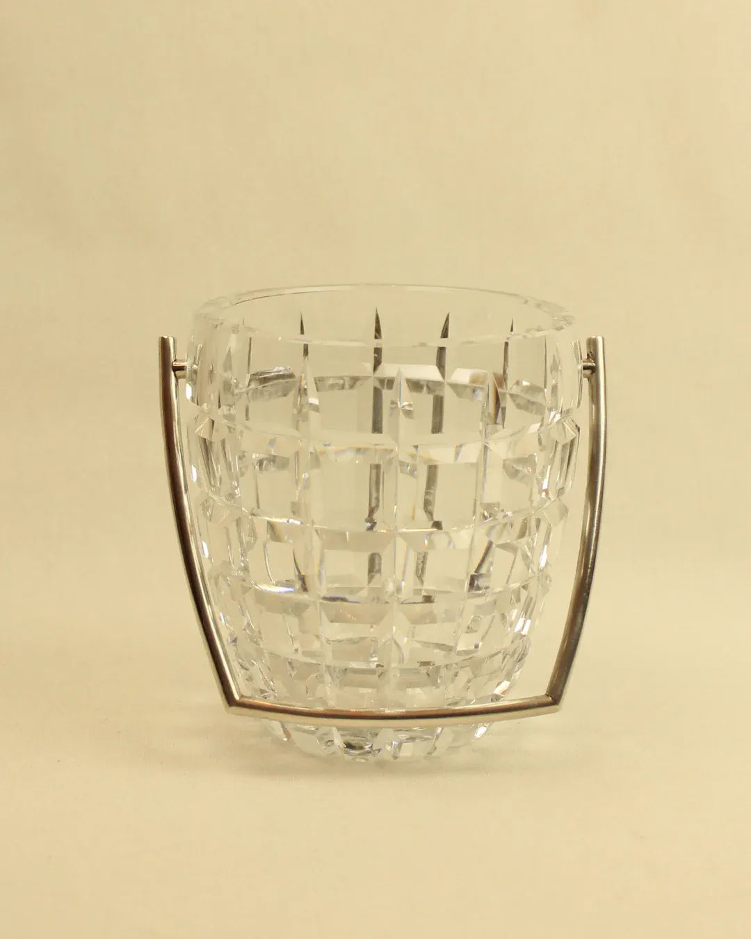 The Crystal Cubitera from Boga Avante Shop sits elegantly against a neutral background. This luxury ice bucket boasts a clear glass construction with a geometric, faceted design and features brass handles on each side, angled to complement the bucket’s distinctive shape. The textured glass reflects light beautifully, creating a captivating shimmering effect.