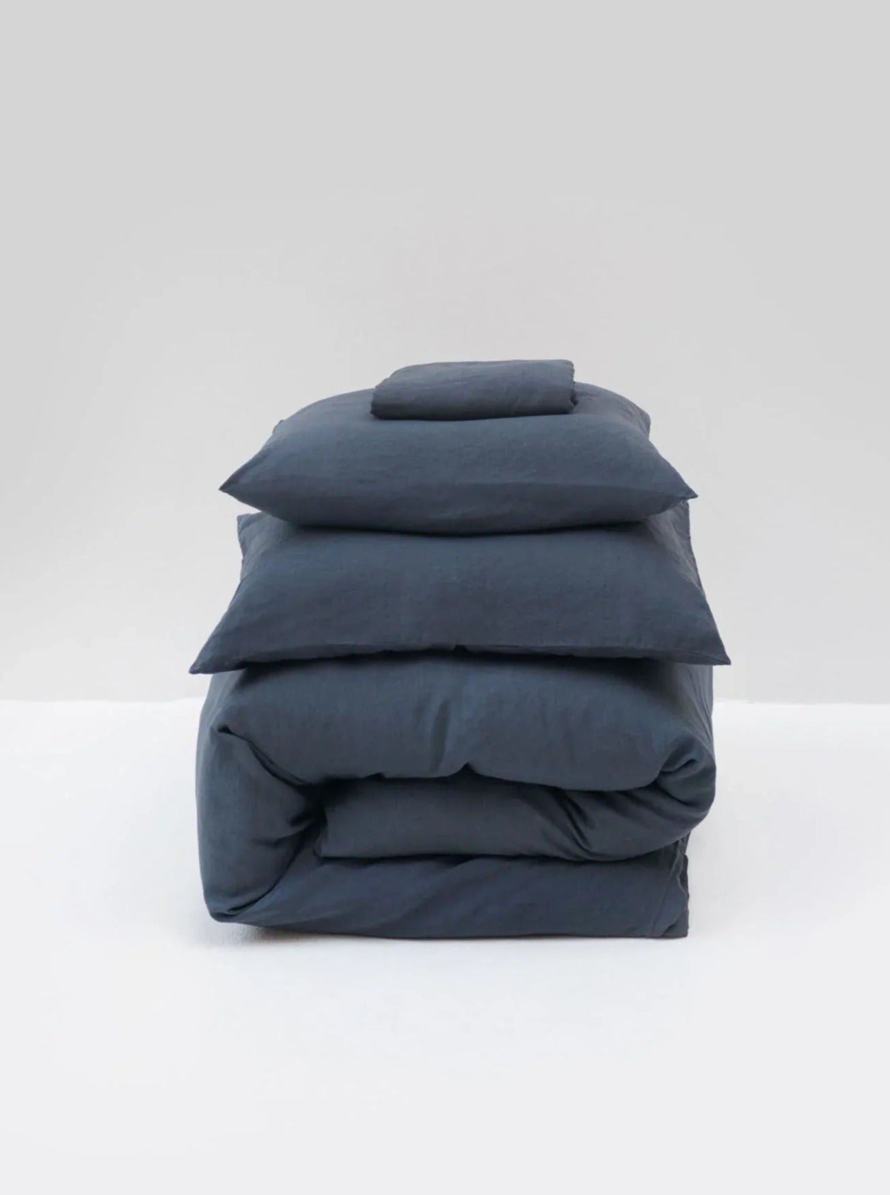 A neatly stacked set of MOST's washed linen bed set in Stormy Blue, including a comforter and four pillows, against a light gray background.