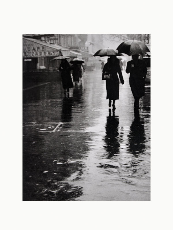 Black and white photo of several people walking with umbrellas on a rainy Paris street, reflected on the wet pavement, creating a moody atmosphere reminiscent of a Maison Plage artwork from "Brassai: Paris & Picasso.