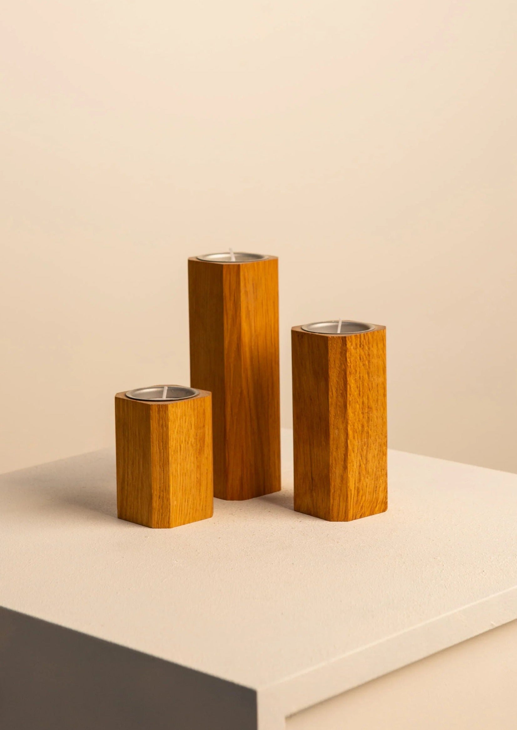 Three Treaptyque oak candleholders of varying heights on a light beige background, each containing an unlit candle, displayed on a soft gray pedestal.