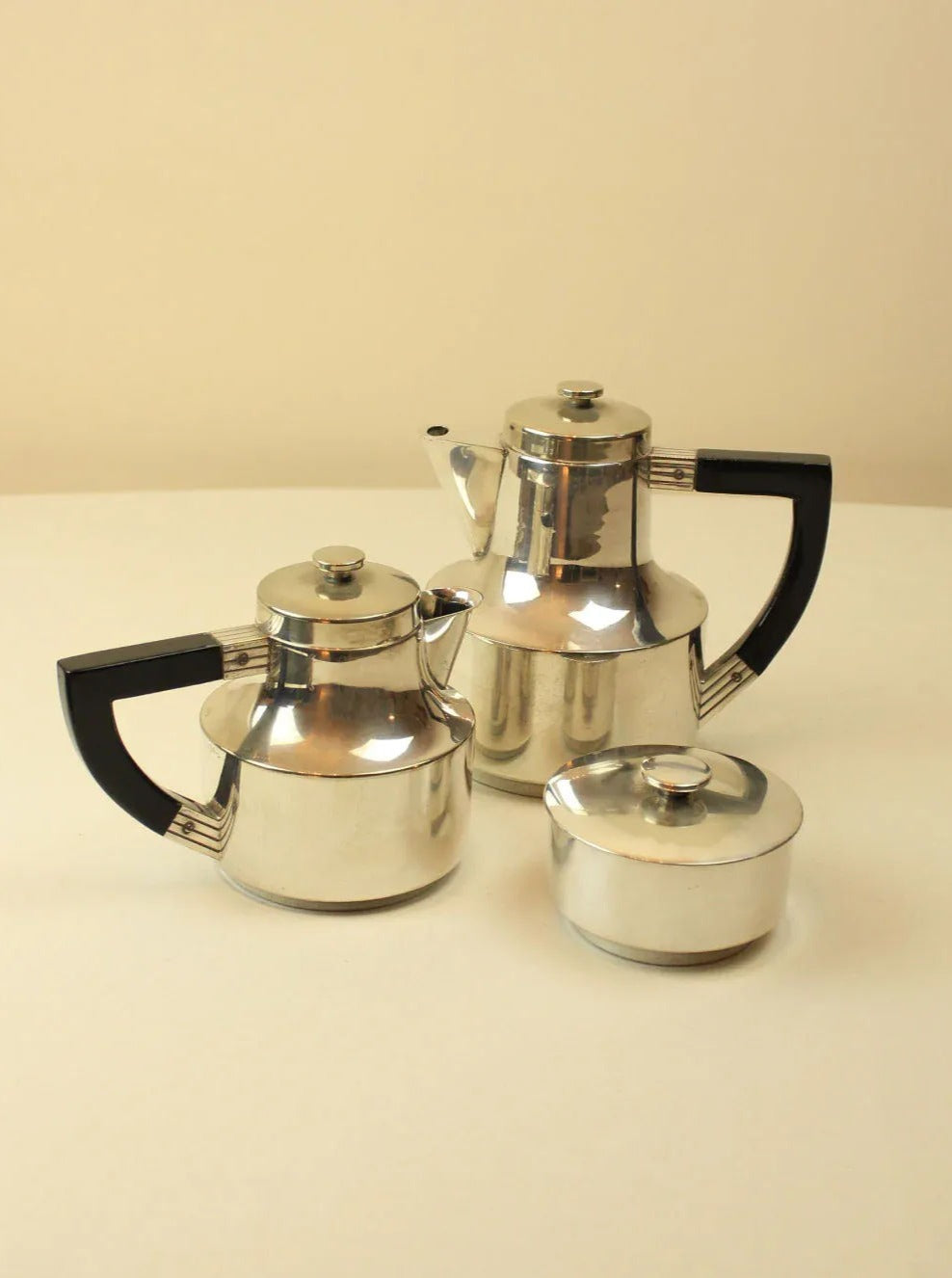 Three vintage stainless steel teapots with black handles on a beige background, ranging in size with the largest having a tall form and the others more rounded, forming part of an elegant Boga Avante Shop Art Deco Coffee and Tea Set.