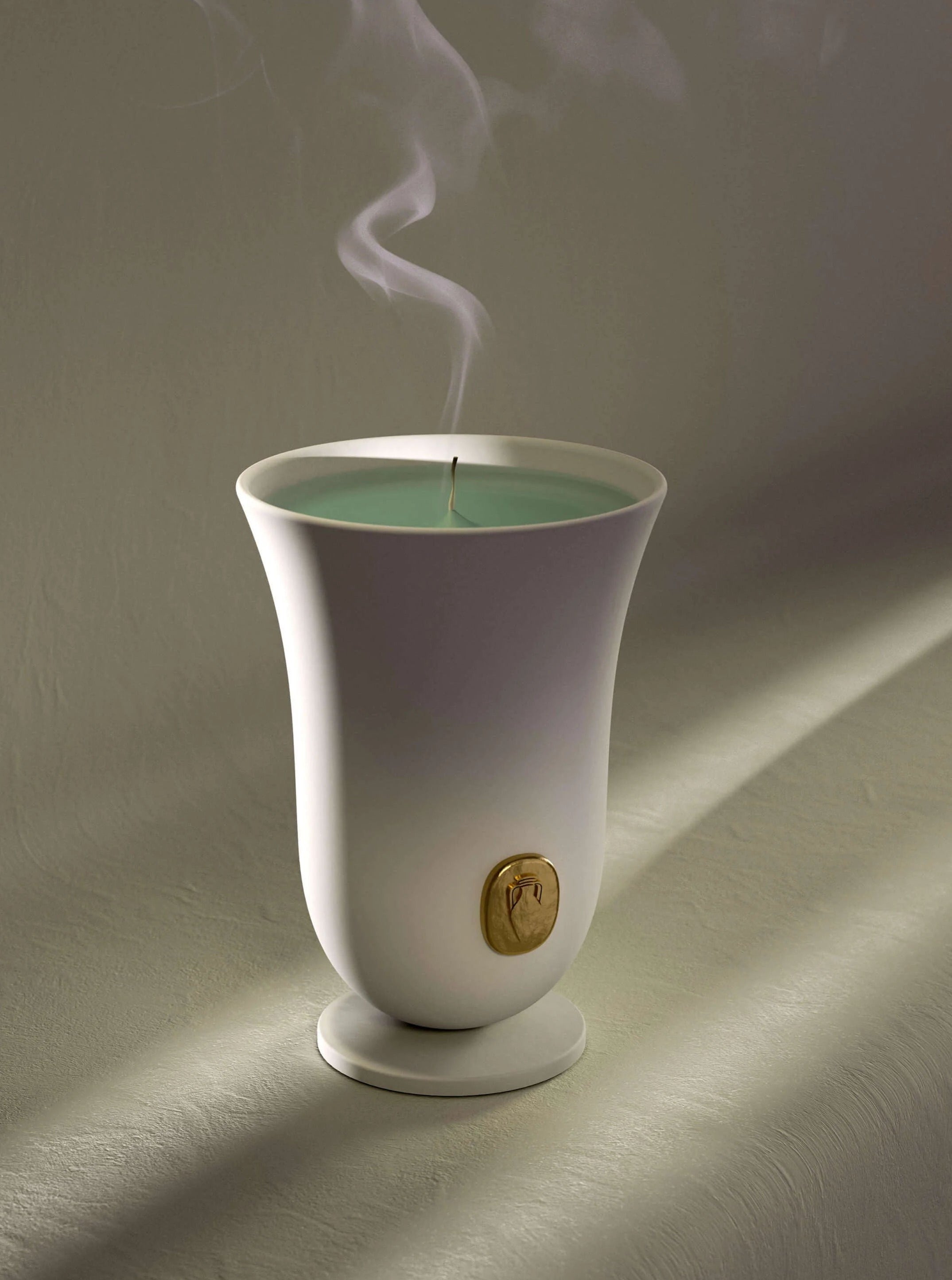 An elegant Bois Vert Candle by LOBJET in a white, tulip-shaped holder with a gold emblem, casting soft shadows on a textured beige surface. The candle's green wax is lit, emitting a thin stream of smoke.