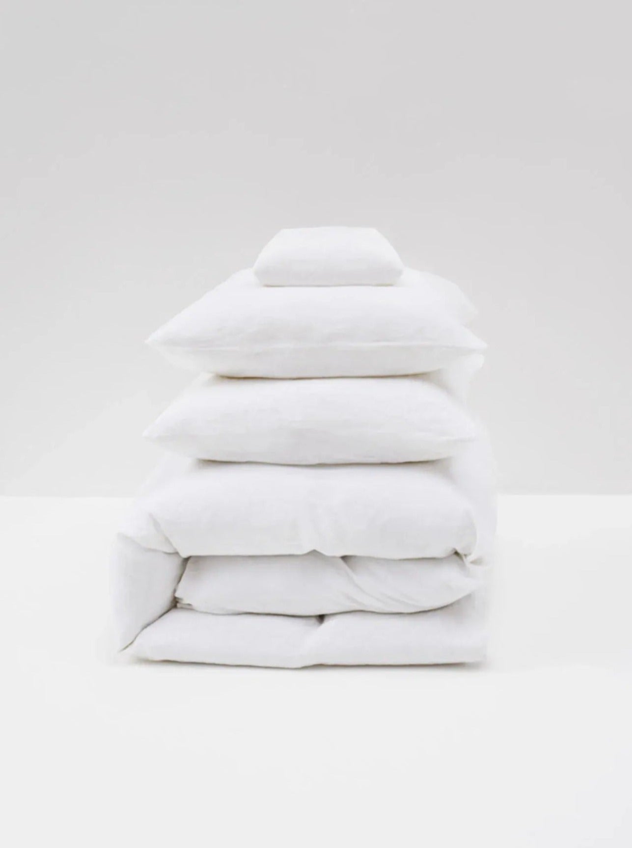 A neatly stacked White Washed Linen Bed Set by MOST, featuring pillows and a folded duvet, is placed against a plain, light gray background, showcasing contemporary design.