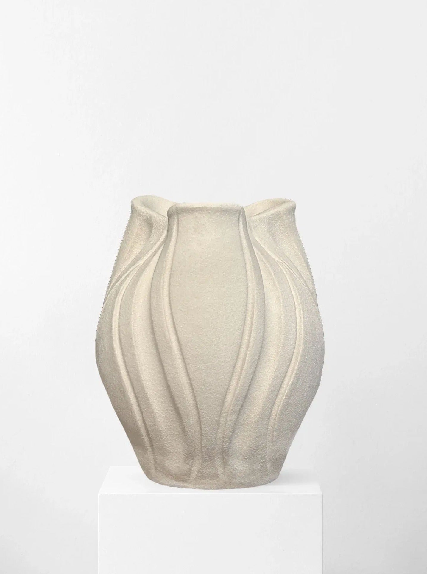 The Sculpt Me Dear Matte Ivory vase by Abomé Studio, displayed on a white pedestal, showcases unique vertical wave-like patterns and a flowing design. This handcrafted beige piece is perfect for adding an organic, sculptural touch to luxury interior decor.