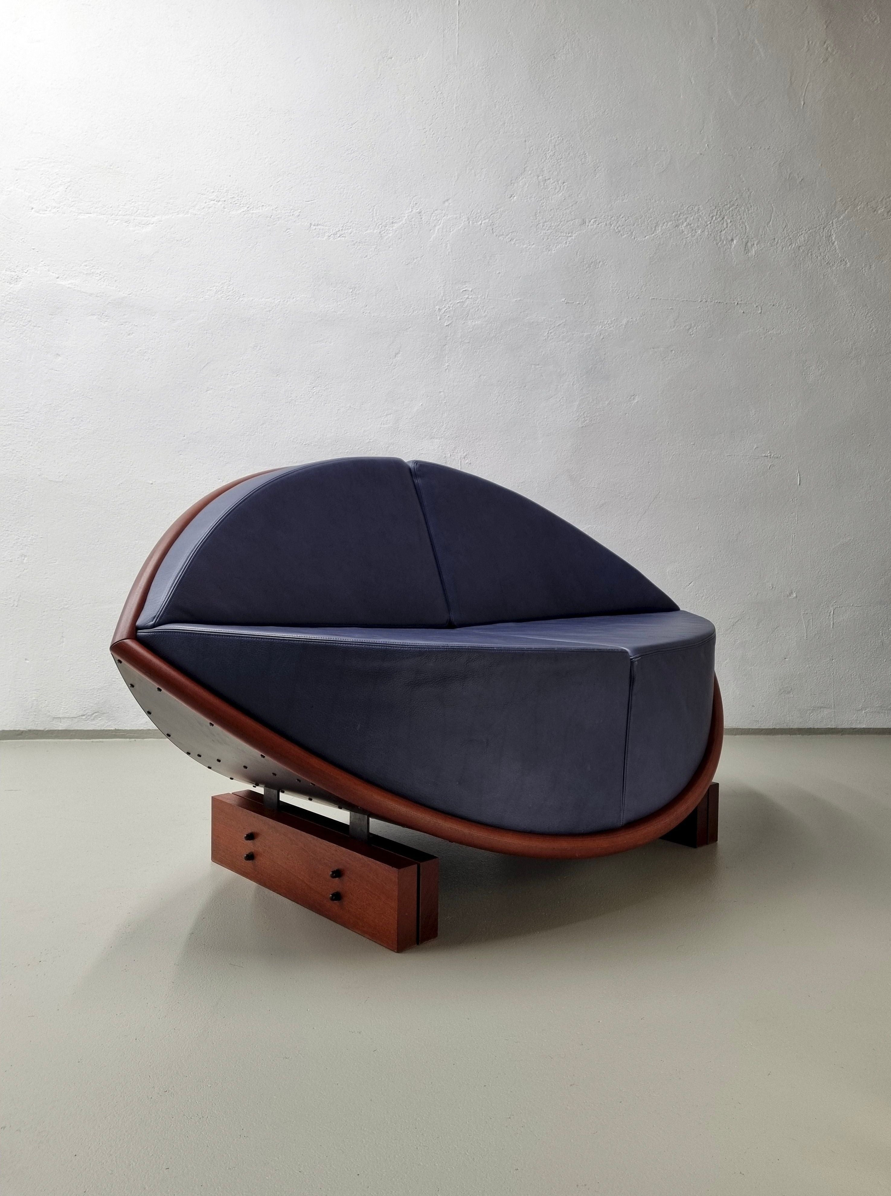 Luxurious oval-shaped chair with a wooden frame, blue cushions, and small storage drawers, designed by Veter Vintage and Sigurdur Gustafsson, displayed against a textured white wall.