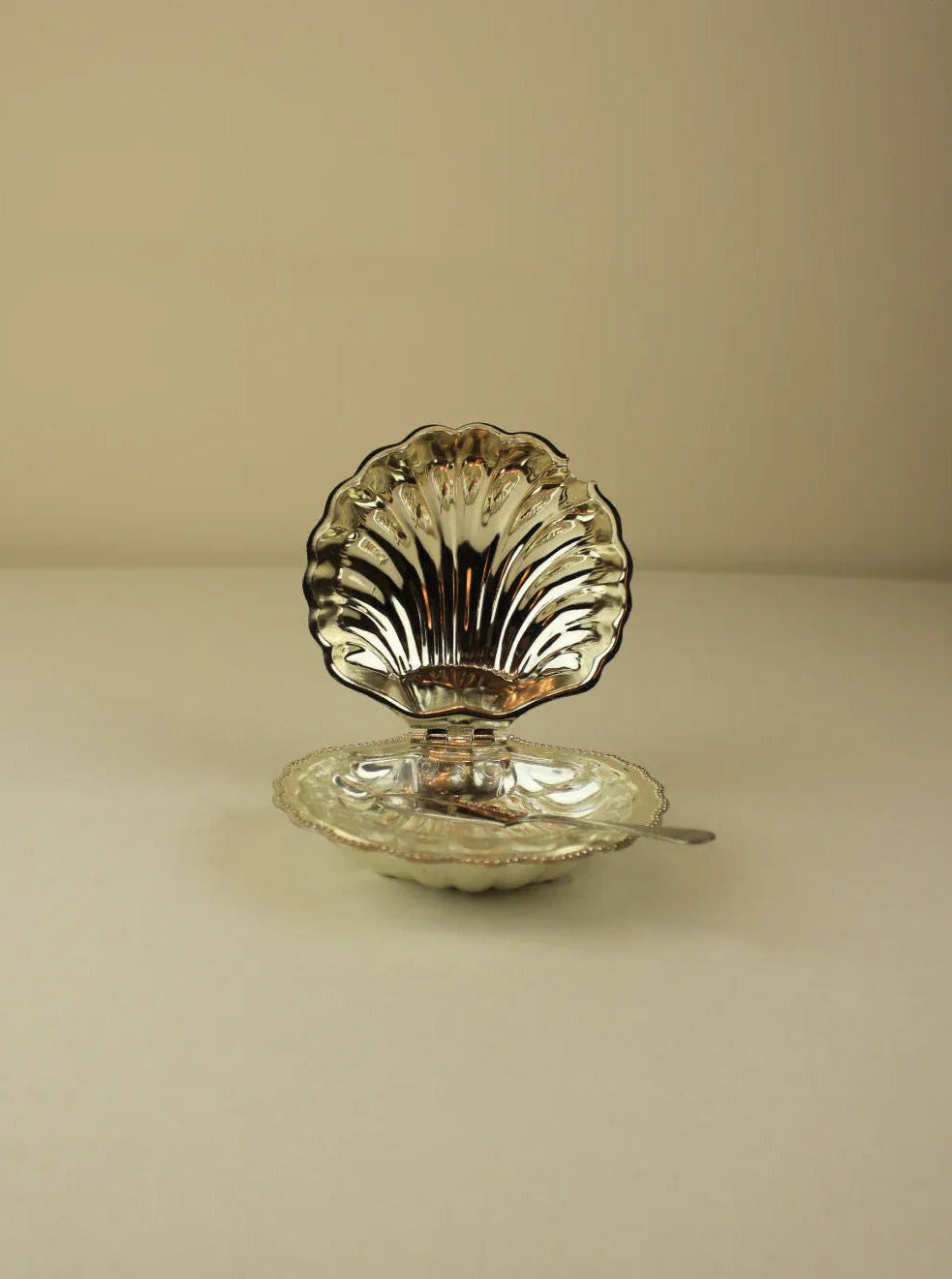 The Shell Shaped Butter Dish from Boga Avante Shop is a vintage silver-plated server set featuring a shell-shaped dish with a matching lid propped open. Included in the set are a small spoon resting on the edge of the dish and a decorative butter knife, all shown against a plain, light-colored surface.