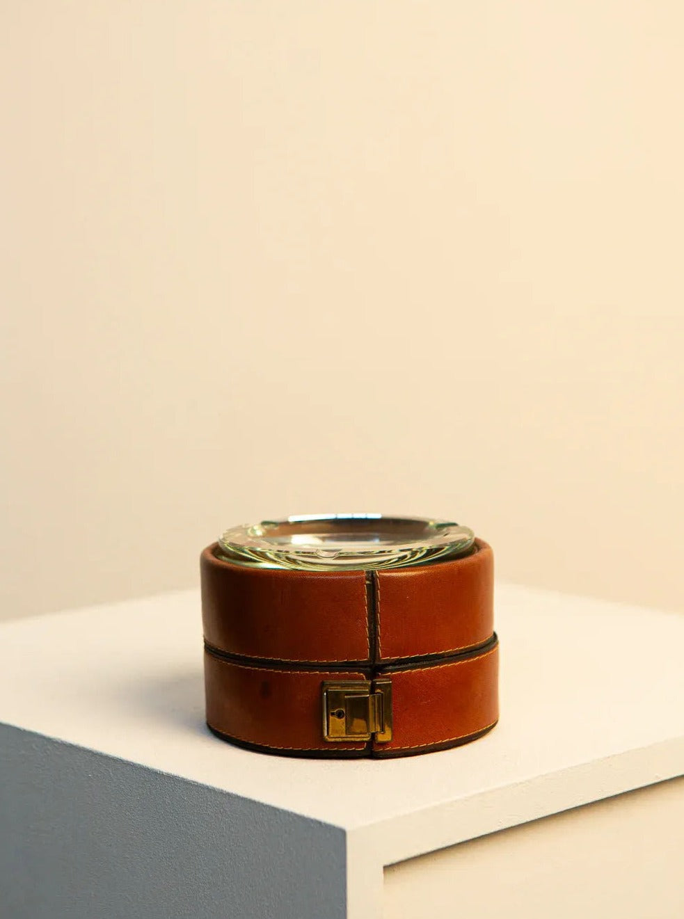 An Italian Ashtray in Brown Leather and 60's Crystal by Treaptyque sits on a white pedestal against a beige background. Reminiscent of 60s craftsmanship, the round brown piece features a removable top, stitching details along the edges, and a brass latch. Its simple and elegant design suggests it's both a storage container and a decorative piece.