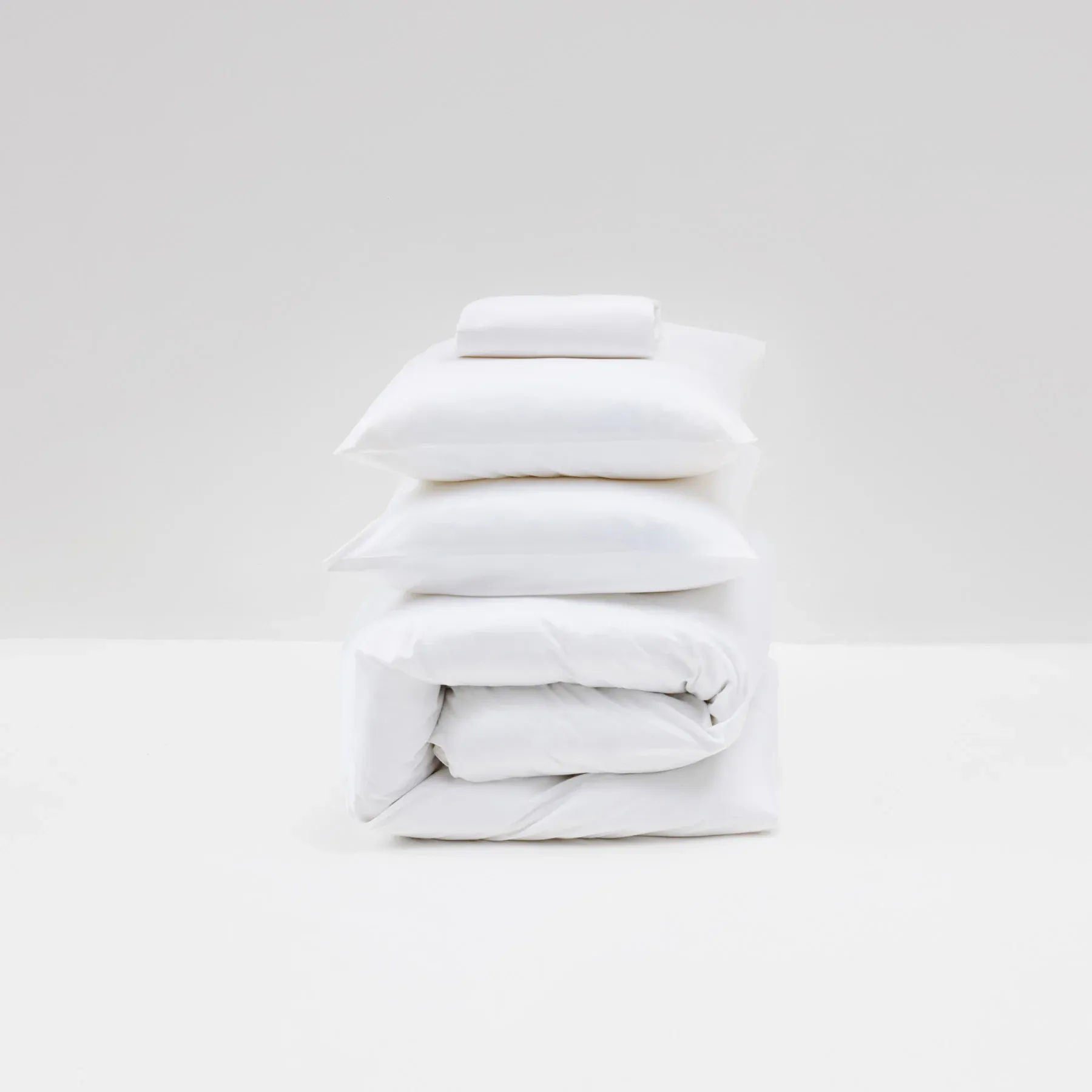 The Cotton Sateen Bed Set in White by MOST showcases contemporary design with two pillows on top, a folded duvet in the middle, and a sheet at the bottom, all neatly stacked against a plain background.