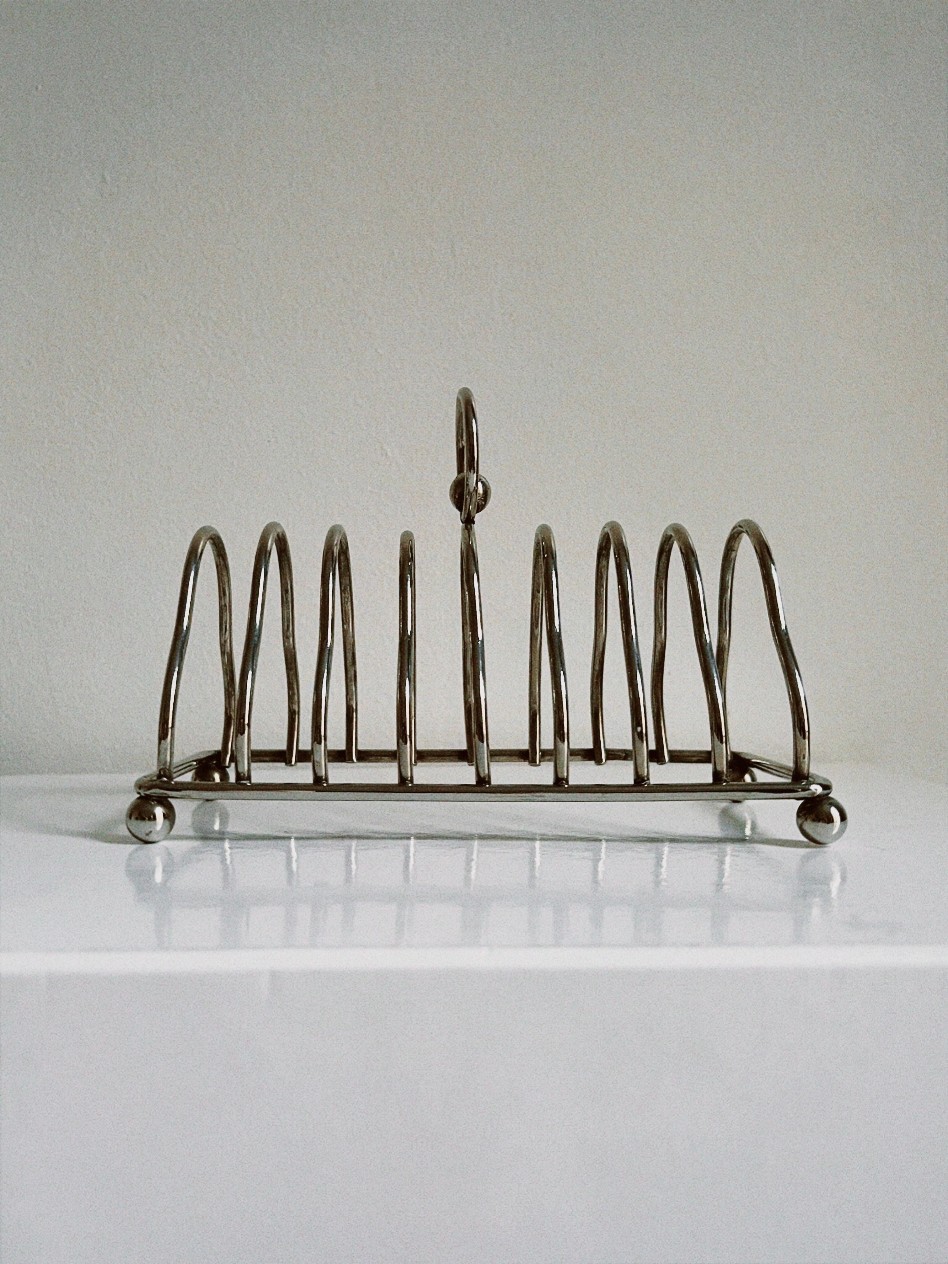 A sleek design Manushi Art Deco Toast Rack with six vertical holders and a central looped handle, placed on a light-colored surface against a neutral background.