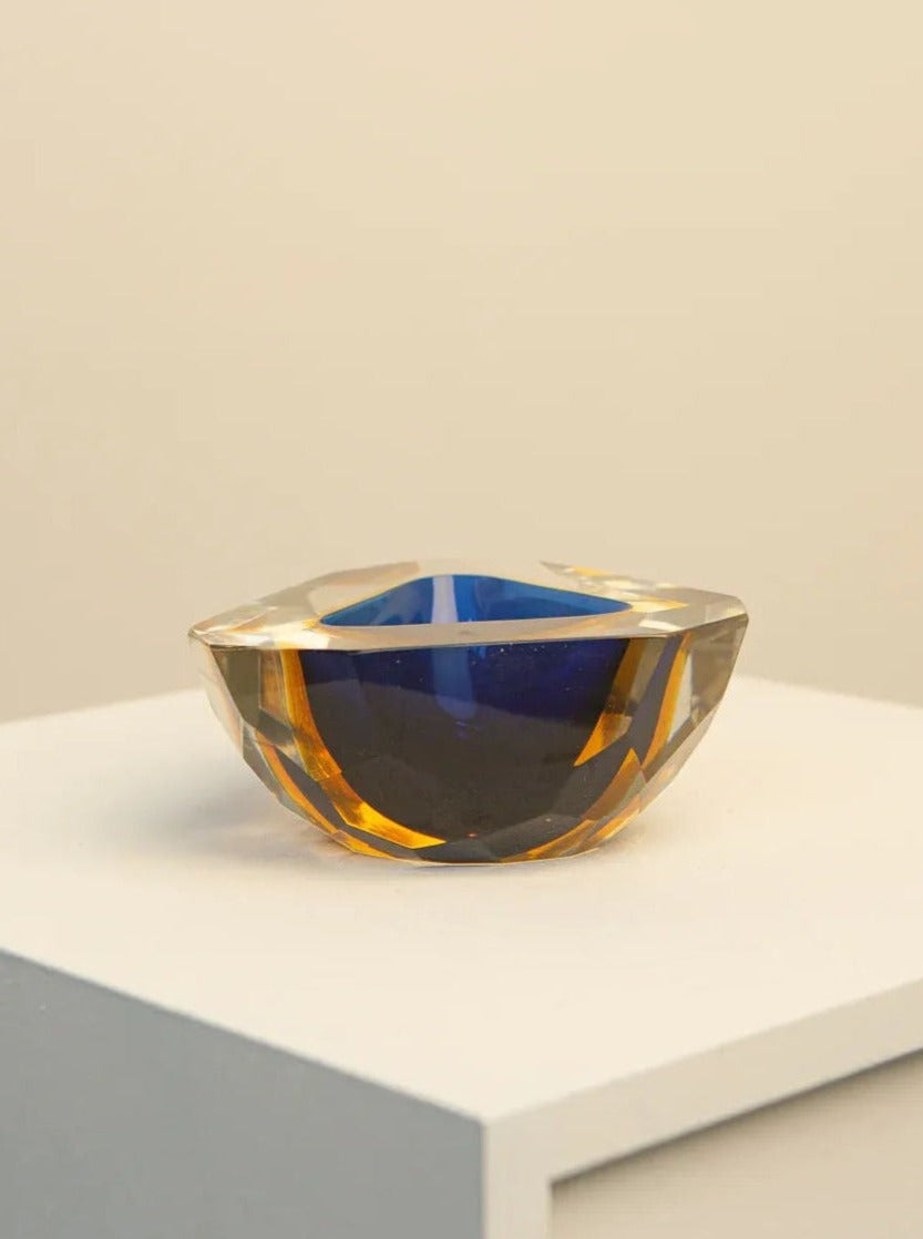 A small, multifaceted Treaptyque Multi-colour Ashtray in Murano Glass by Flavio Poli 60's sits on a white surface. The ashtray is primarily blue with amber-toned sections, and its geometric design creates a striking visual effect. Reminiscent of Flavio Poli's work, the background is a simple, light beige wall that emphasizes the ashtray's colors.