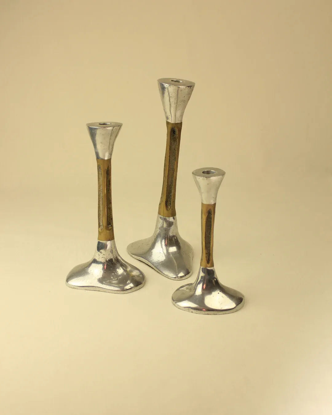 A Trio of Brutalist Candleholders from Boga Avante Shop, made of silver metal with brown accents and varying in height, are arranged together on a light beige background. These 1980s candle holders feature unique brutalist designs with wide bases and narrow tops, making them an intriguing addition to home decor.