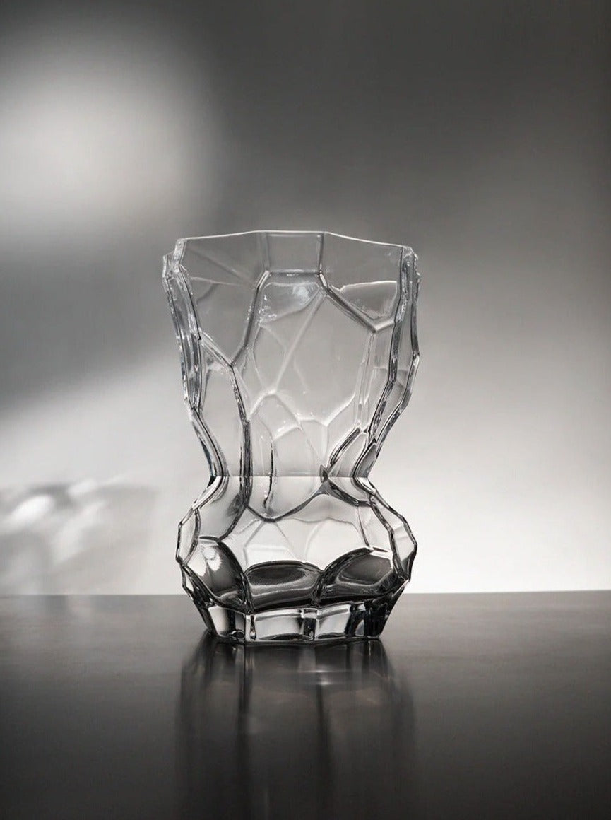 A Reflection Vase - Clear by Hein Studio sits on a reflective surface against a soft gradient gray background. The vase features a faceted design that mimics a crystal structure.