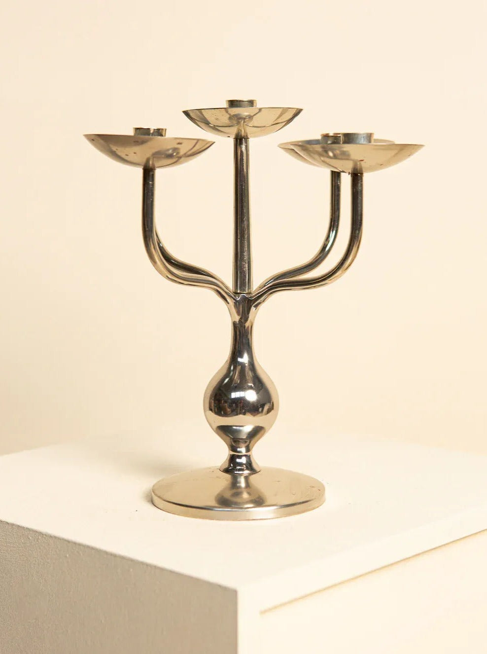 A sleek, three-armed silver-plated candleholder with an elegant design from the 70s Italian "Medusa" collection by Treaptyque, placed on a white pedestal against a beige background. Reminiscent of 80s interior design, each arm curves upward to hold a candle socket, creating a symmetrical and minimalistic appearance.