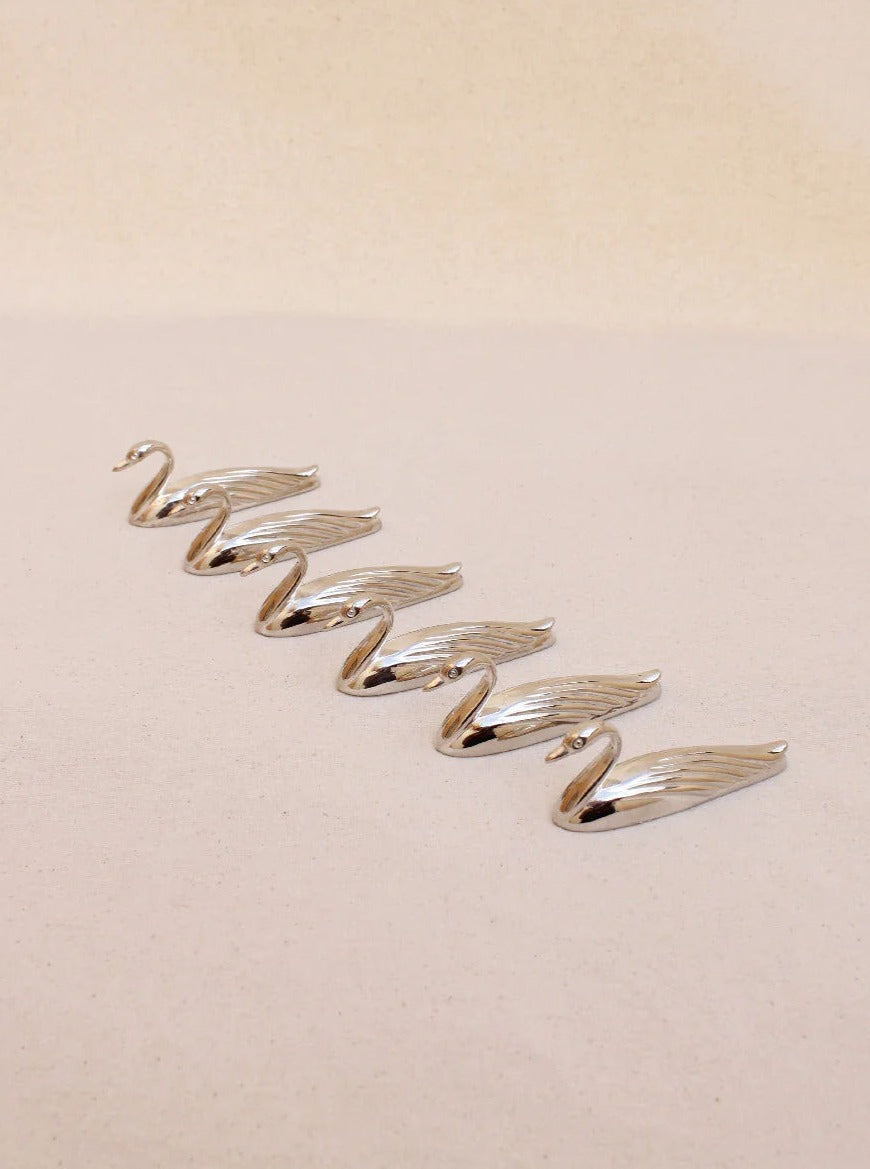 A set of six Boga Avante Shop's handcrafted silver swan cutlery holders is elegantly arranged on a light beige surface, each featuring gracefully extended wings and lifted heads, highlighting their sculptural beauty against the minimalist backdrop.