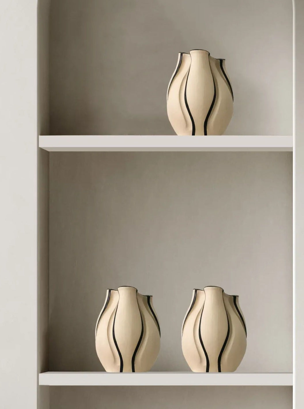 Three "Sculpt Me Dear Black Lines" vases by Abomé Studio, featuring beige tones and black wavy lines, are elegantly showcased on two minimalist white shelves against a light gray wall, reflecting contemporary design.