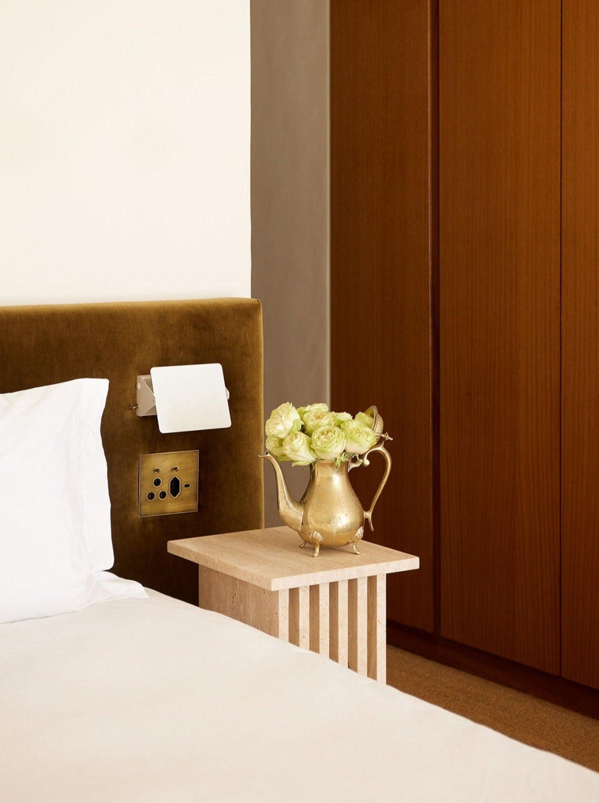 A cozy bedroom corner featuring a plush olive green headboard, a Lemon Vondel Side Table in Travertine with a vintage silver pitcher filled with light green roses, and white bedding.