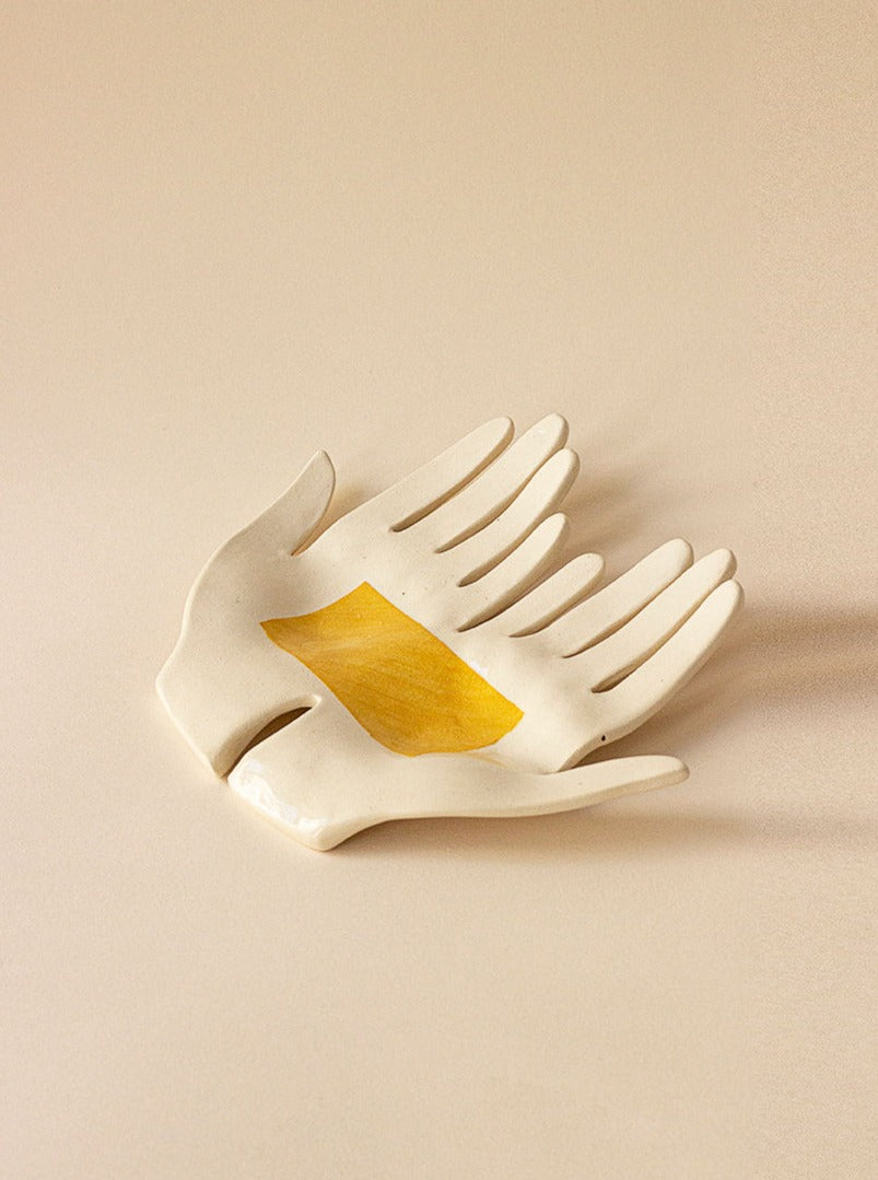 An artistic handmade Main Edition stoneware sculpture depicting two overlapping hands, one with a golden yellow section in the palm, set against a neutral beige background.