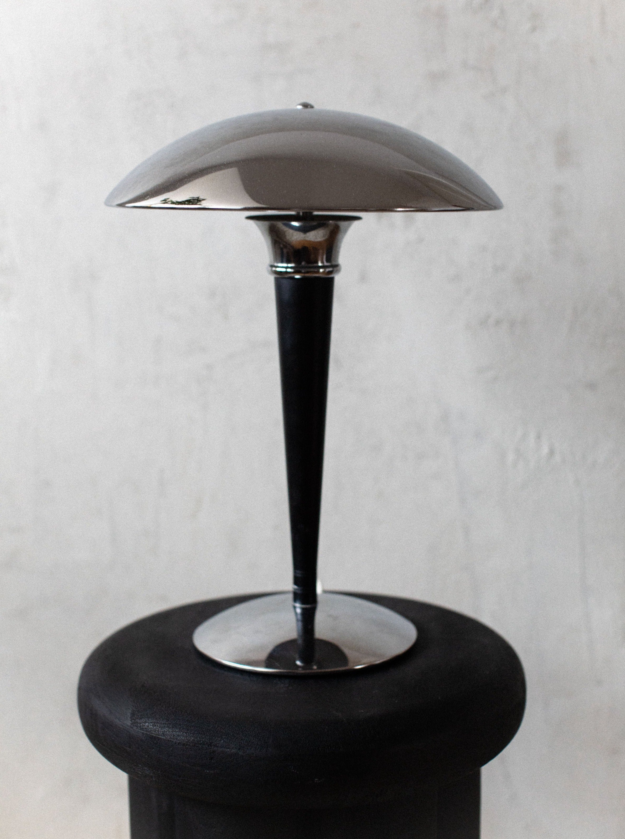 A sleek, modern lamp influenced by Bauhaus principles with a metallic dome-shaped shade and slim black stem, standing on a round black base against a textured white wall. This is the Out For Lunch Bauhaus Table Lamp.