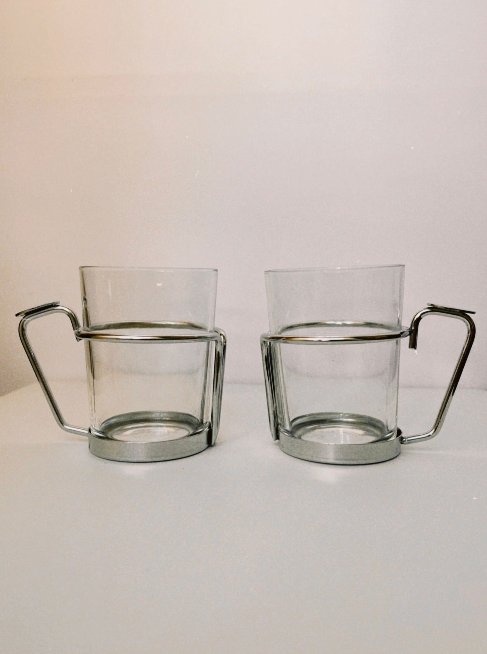 Two clear Manushi French tea glasses with stainless steel holders, placed side by side on a white surface against a plain background.