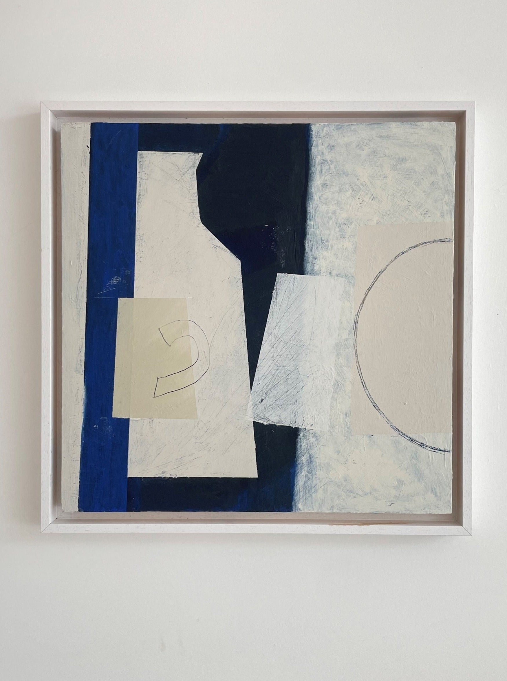 A framed Blue Still Life I abstract acrylic painting on a wall, featuring geometric shapes in shades of blue, white, and a touch of beige by Lara Voce.