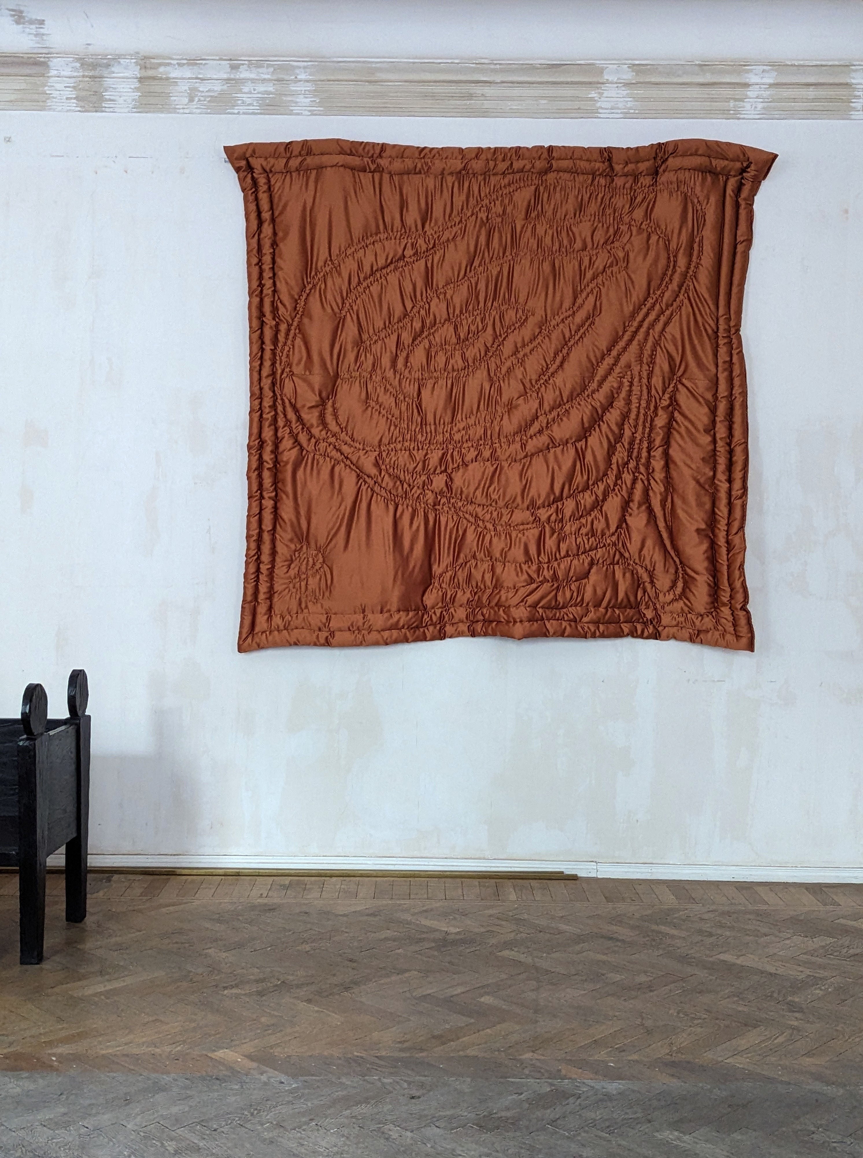 A large, crinkled Dowry Blanket 2 by Rooms Studio hung on a white wall in a room with wooden floors and a minimal black chair to the left.