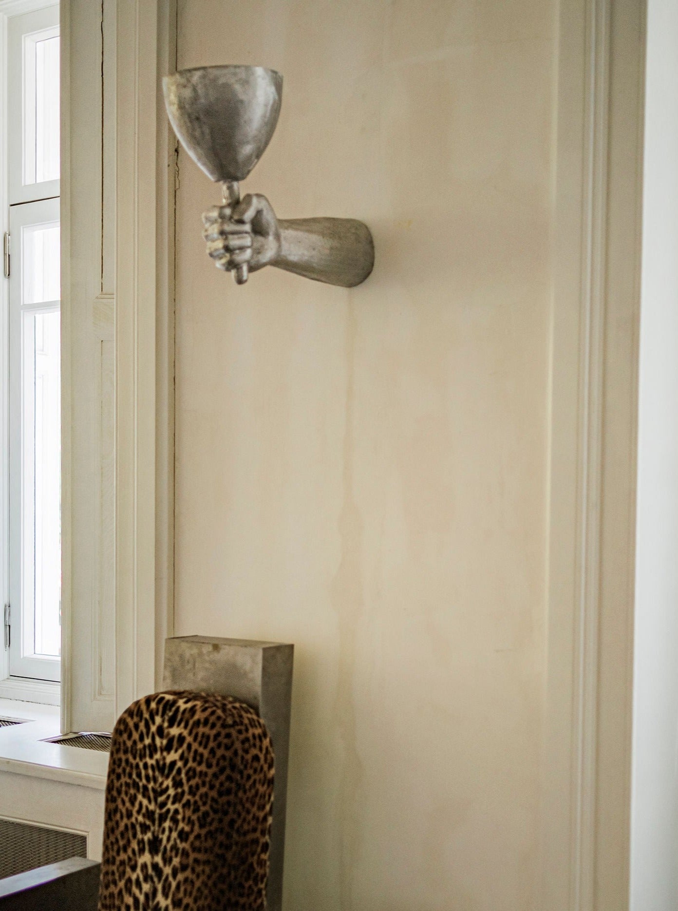 A sculptural Dracula Wall Scone Aluminium shaped like a hand holding a cup, crafted from high-quality materials, mounted on a pale wall beside a window with white frames and a leopard print chair partly visible. Designed by Rooms Studio.