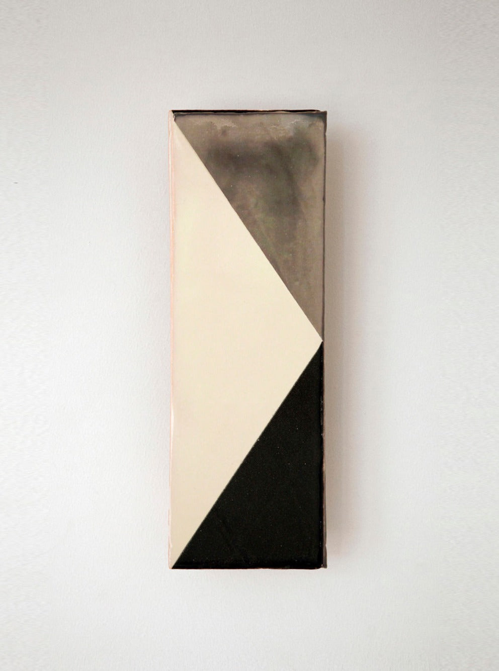 A vertical, narrow artwork on a wall featuring a diagonal division with one half painted in black and the other in white, crafted using Violaine d'Harcourt handmade tiles.