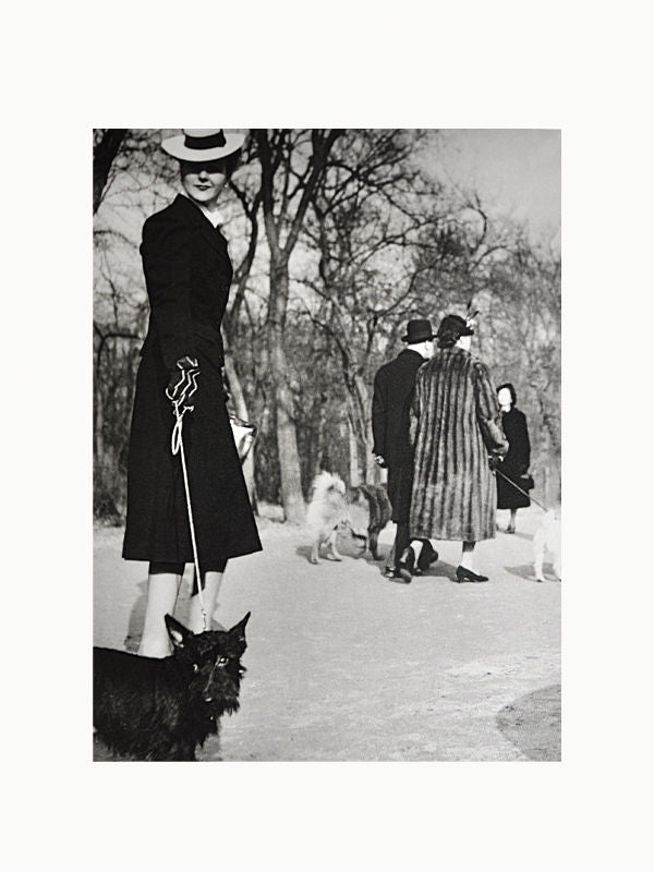 A monochrome photo of an elegant woman in a coat and wide-brimmed hat seen from behind, holding a leash attached to a cat in the foreground, with people and other animals blurred in the background by Maison Plage's Brassai: Paris & Picasso.