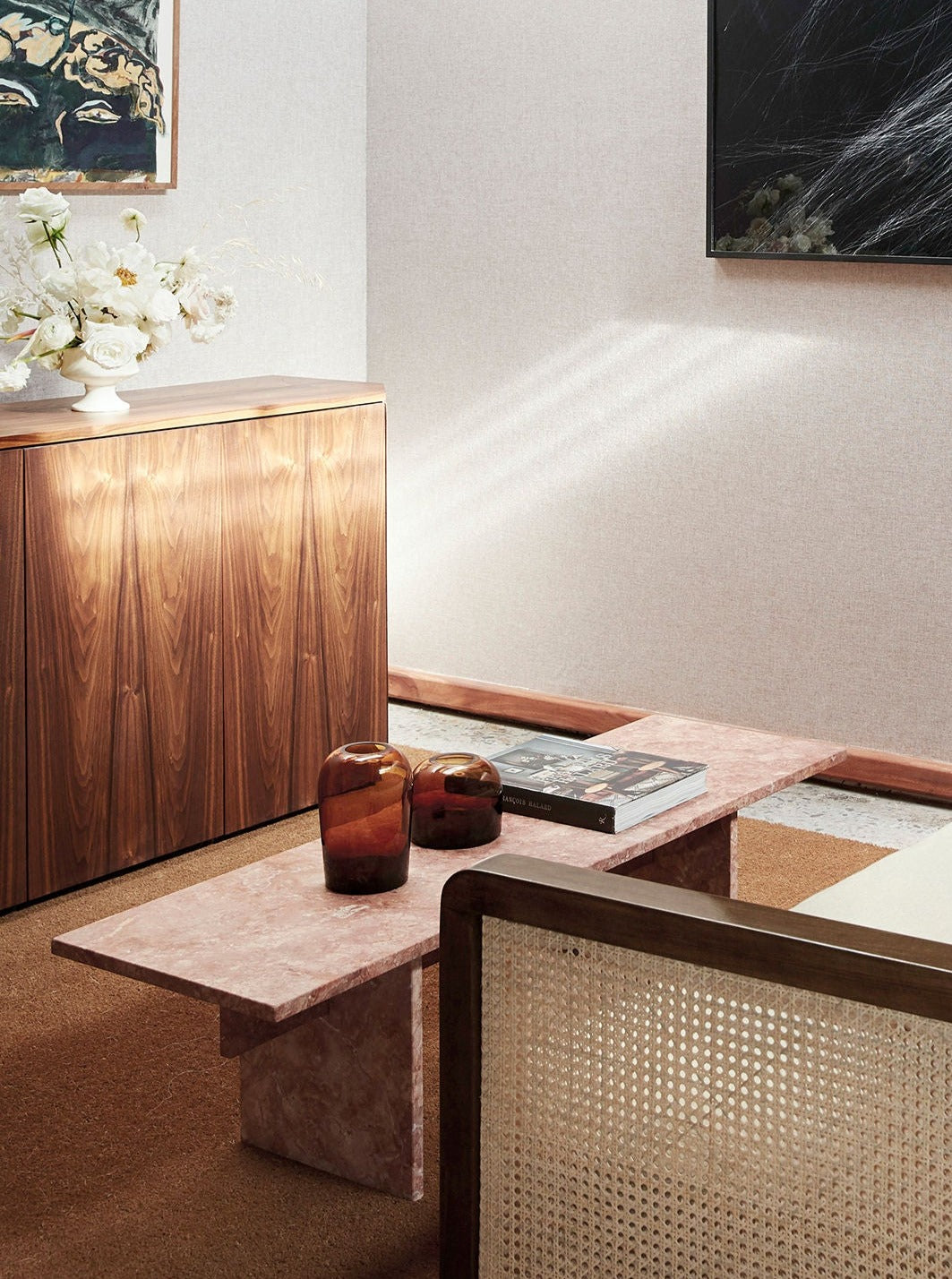 A contemporary living room featuring a Lemon Vondel Coffee Table in Red Travertine, wooden cabinet, and a cane chair. The decor is complemented with a vase of white flowers and abstract wall art.