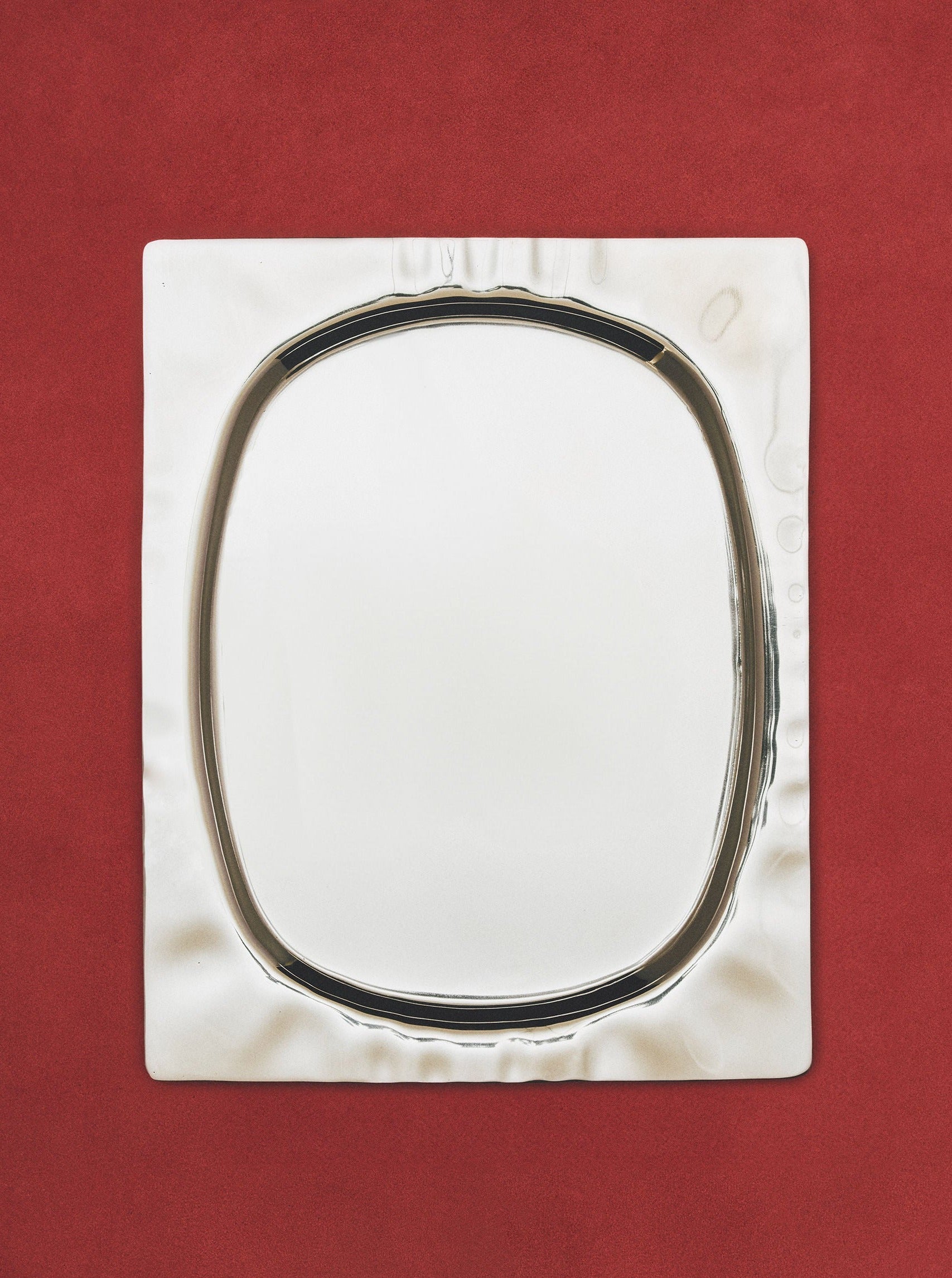 A square silver tray with a polished finish and a reflective, oval-shaped center placed on a deep red background with a textured surface. (Product Name: 104-1 by Ananas Ananas)