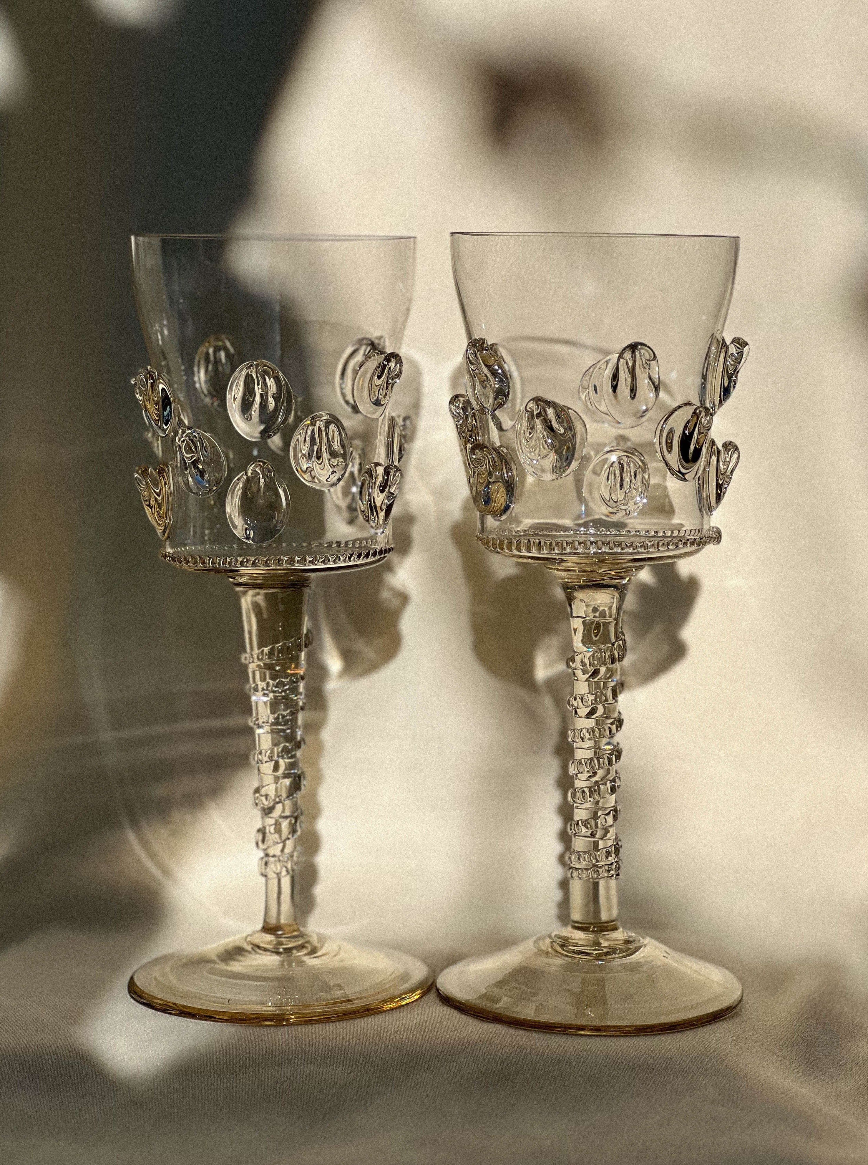Two ornate, crystal wine glasses featuring detailed, raised bubble-like designs on the upper portion and intricately textured stems. Mouthblown using Bohemian glassmaking techniques, the Pair of Mouthblown Wine Glass - Dots by Lucie Claudia Podrabska are placed side by side against a softly-lit background, creating delicate shadows.