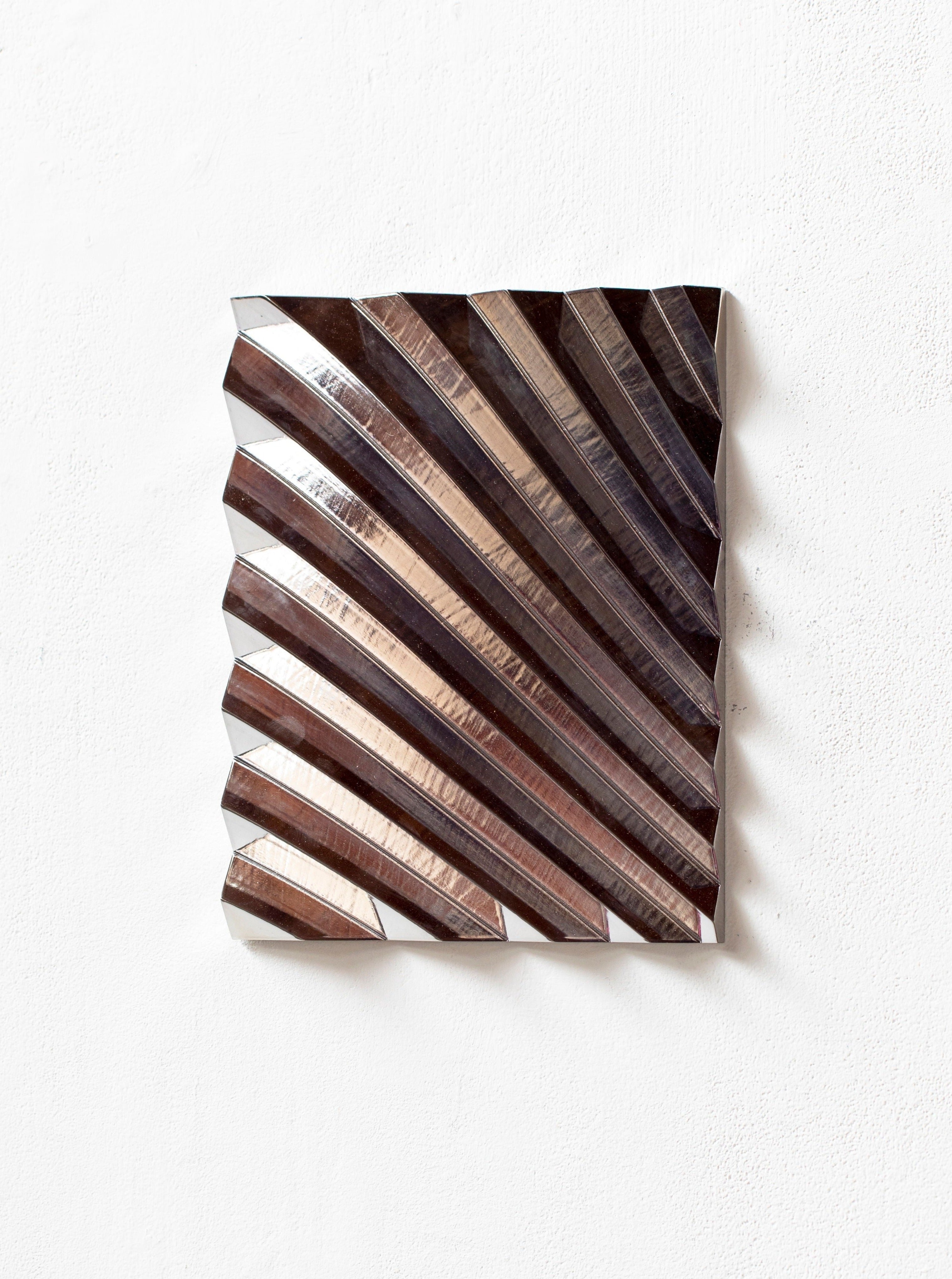 The "Wave Chrome Silver" by Studio Arno Hoogland is a sculptural metal artwork with a square design featuring diagonal, raised grooves that create a geometric pattern. Its metallic finish reflects light, enhancing texture and offering a dynamic, three-dimensional look on a plain white background.