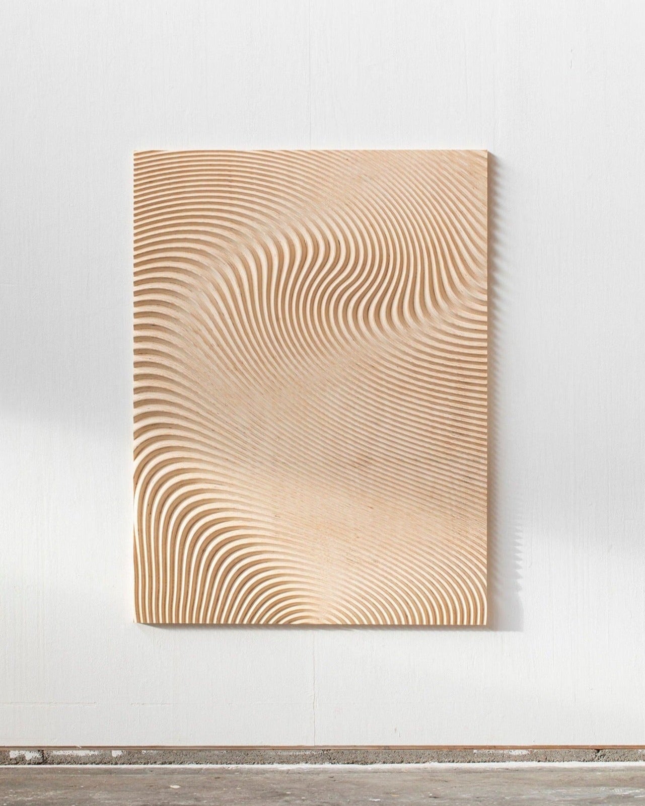 A Wave Naturel L wooden wall panel with undulating, concentric lines, creating a three-dimensional wave pattern, displayed against a white wall in a minimalist setting by Studio Arno Hoogland.
