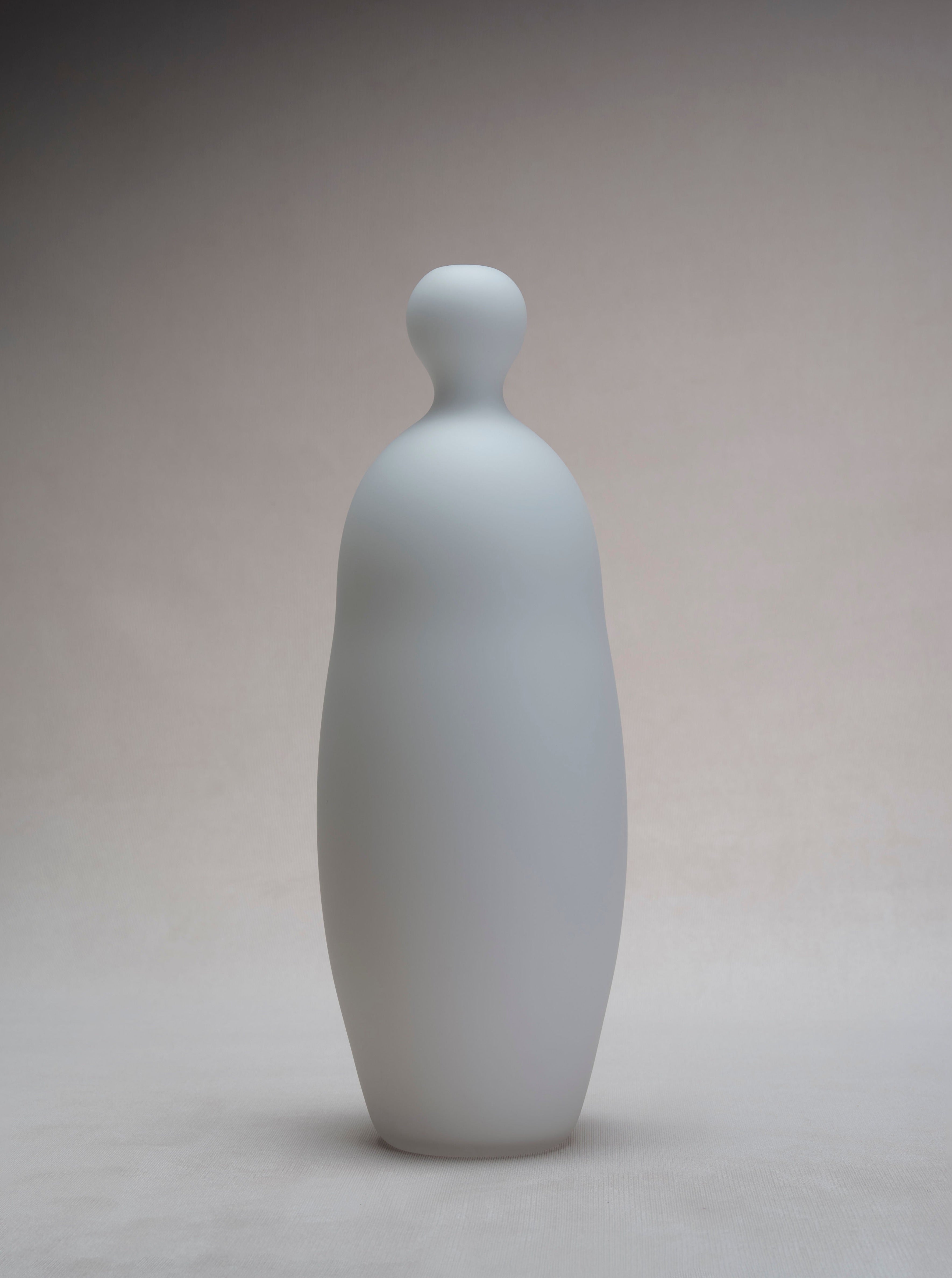 A minimalist Verre d'Onge white ceramic sculpture resembling a stylized human figure with a smooth, curved form set against a soft gray background, crafted by Montreal artisans.