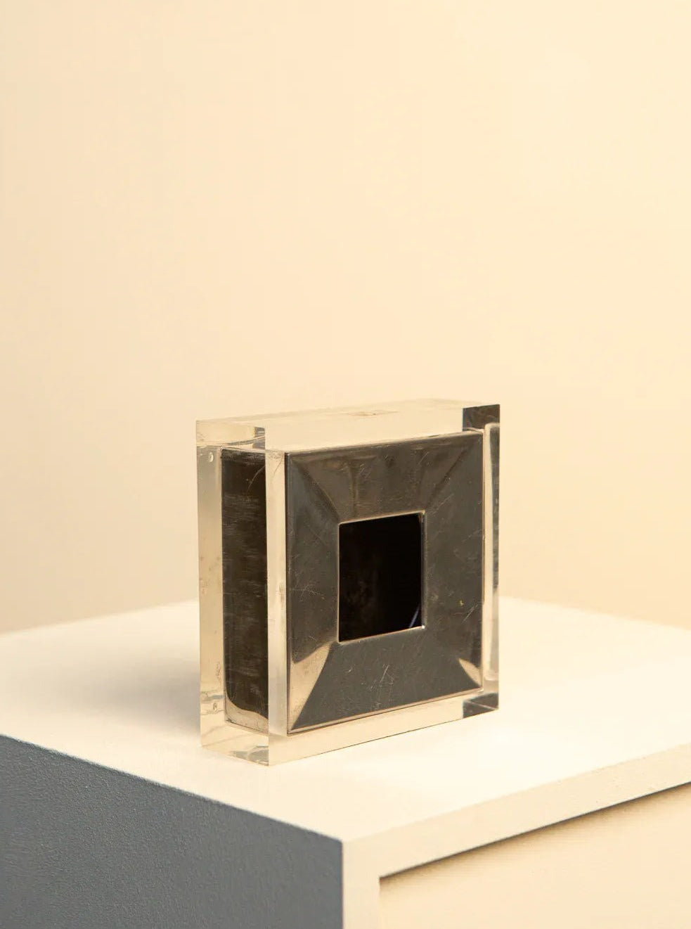 A small, transparent, rectangular object with a dark square cutout in the center is placed on a light-colored surface against a cream-colored background. This minimalist piece exudes Italian design elegance by Treaptyque and could easily be mistaken for the Metal and Plexiglas Ashtray by Guzzini from the 70's.