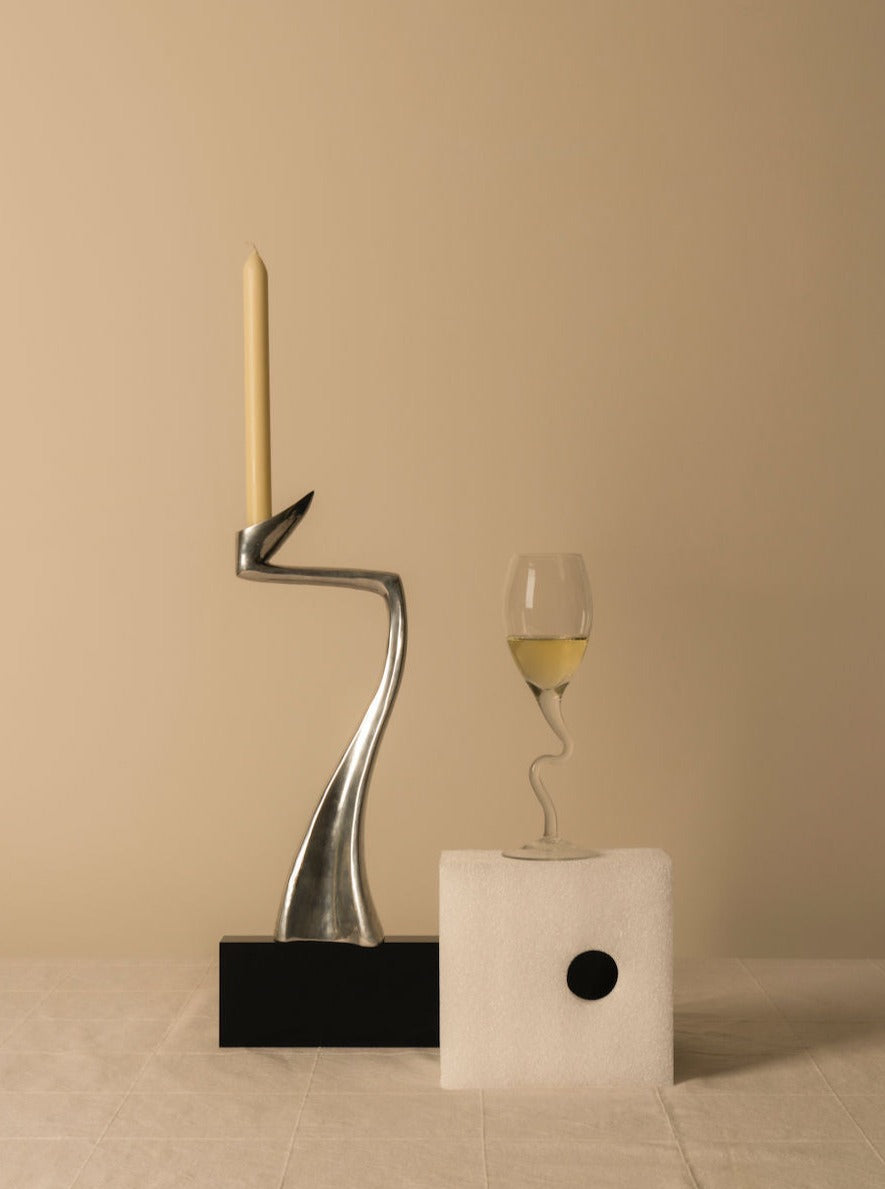 On a beige background, the Vintage Matthew Hilton Candle Holder by Les Objets Amsterdam with a lit candle adorns the left. Next to it, a wine glass of white wine rests on a textured white block with a small black hole, embodying minimalist aesthetic design.