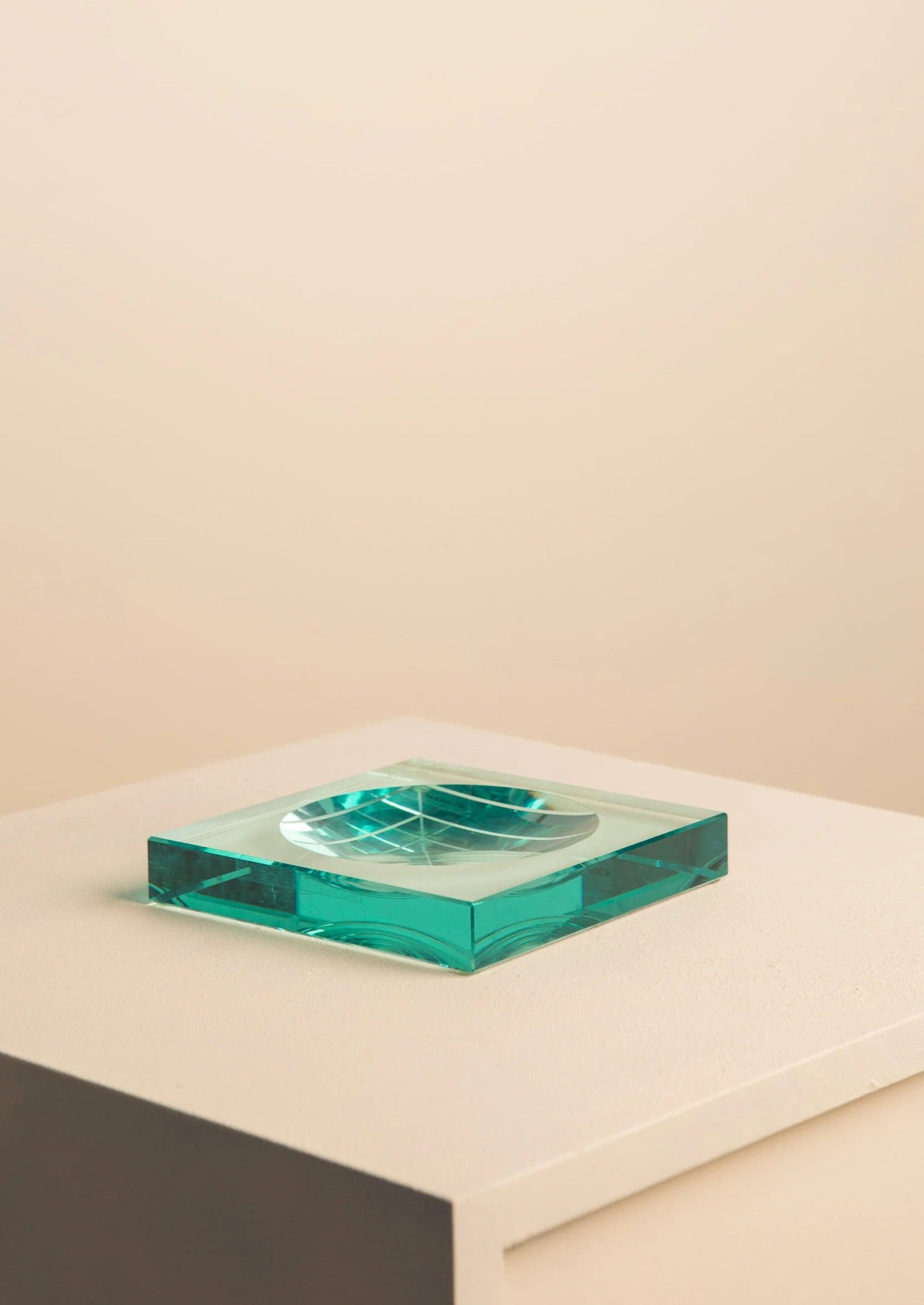 The Treaptyque Glass Vide Poche by Fontana Arte, a sculptural square green glass block with a circular pattern, sits on a white pedestal against a beige background. Its handcrafted transparent edges catch the light, creating vibrant contrasts with the muted setting.