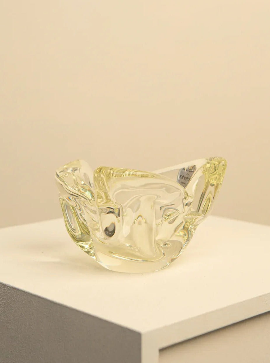 A sleek, modern decorative piece, this Lemon Yellow Crystal Bowl by Sèvres 60's from Treaptyque features clear, abstract lines with curvy, flowing edges. Displayed on a white pedestal against a plain beige background, the bowl's intricate design showcases reflections and refractions, giving it a shimmering appearance.