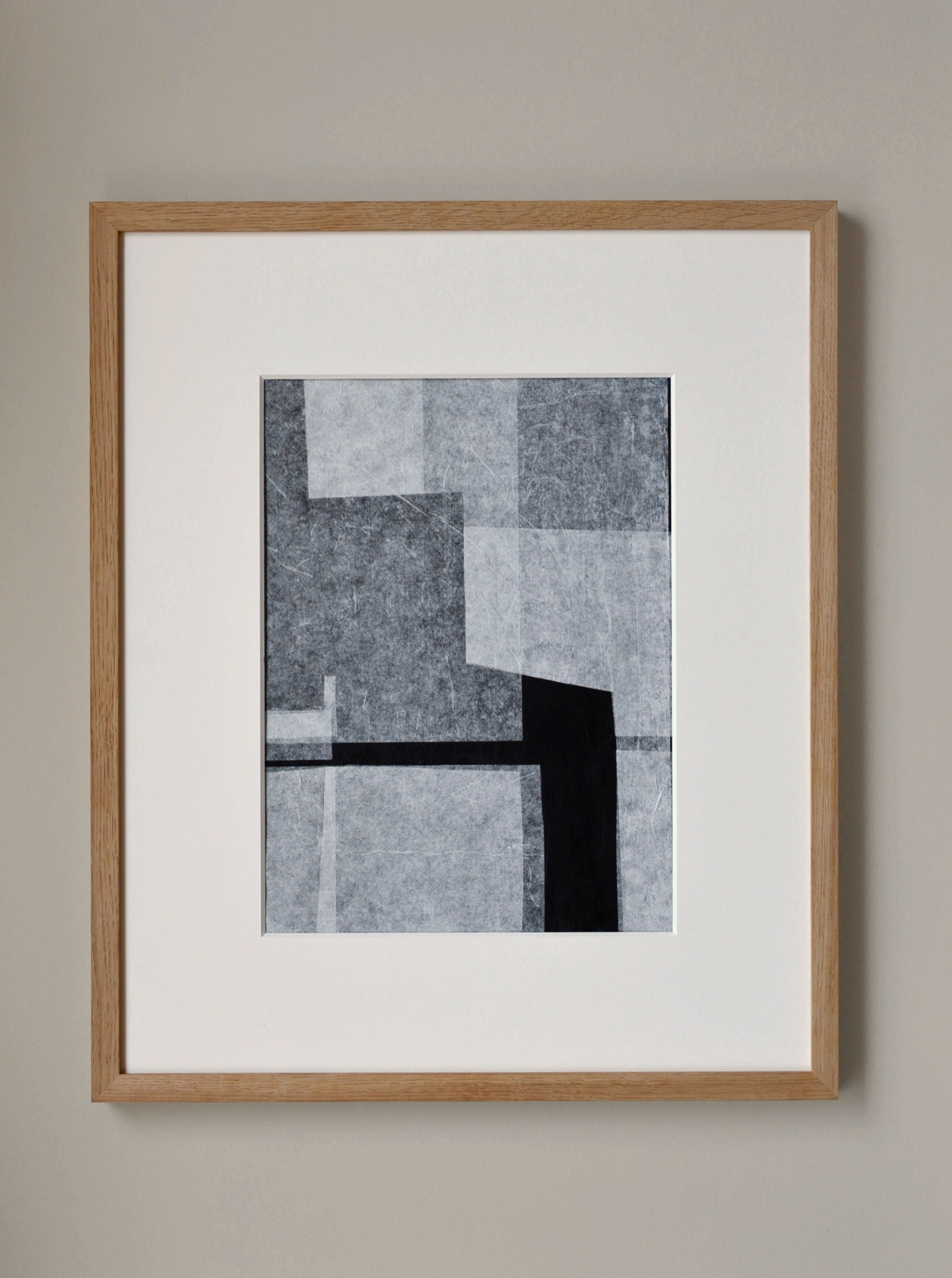 Abstract black and white Stone and Gap No.05/22 paper collage artwork in a light wooden frame, mounted on a light gray wall. The art features overlapping rectangles and squares in various shades of gray and black by Julita Elbe.