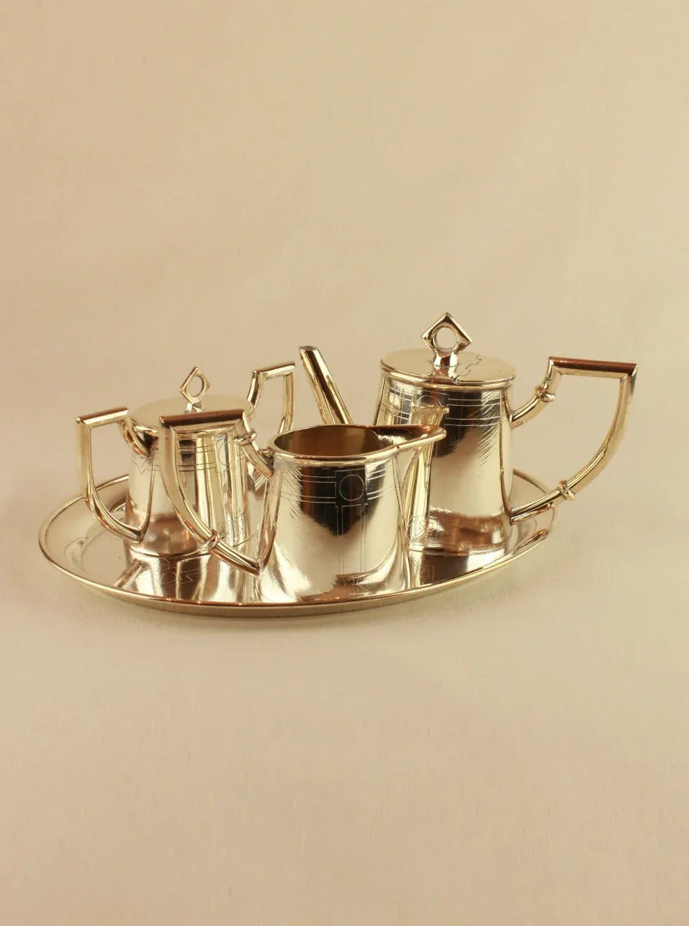 A polished silver Art Deco Tea Set by Boga Avante Shop on a tray, including a teapot, sugar bowl, creamer, and a waste bowl, all with matching lids and elegant handles, against a beige background.