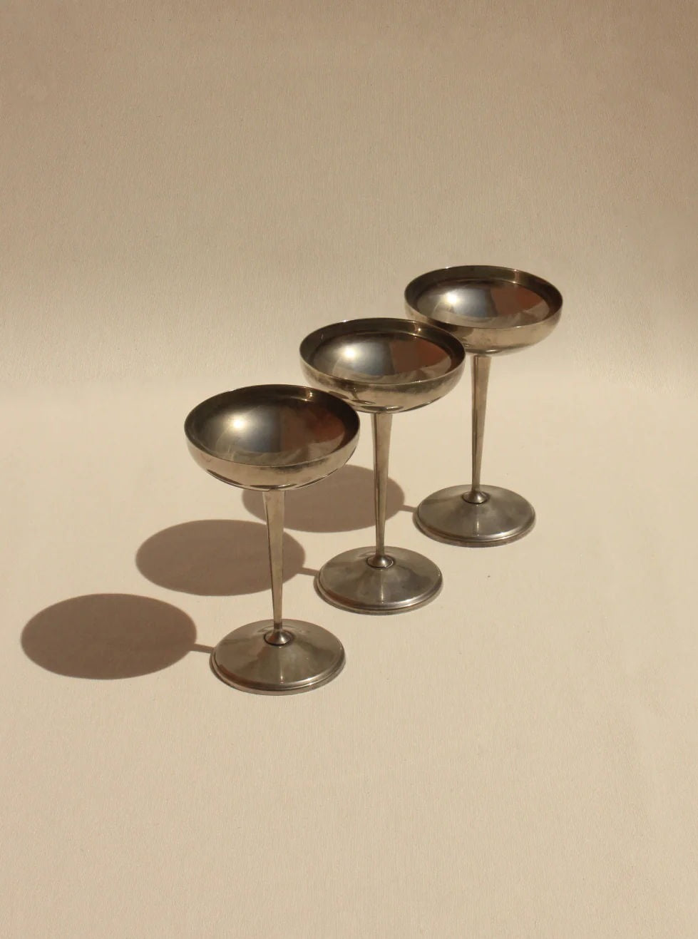 Three metallic Boga Avante Shop cups arranged in a triangular formation on a beige surface, creating soft shadows under warm lighting.