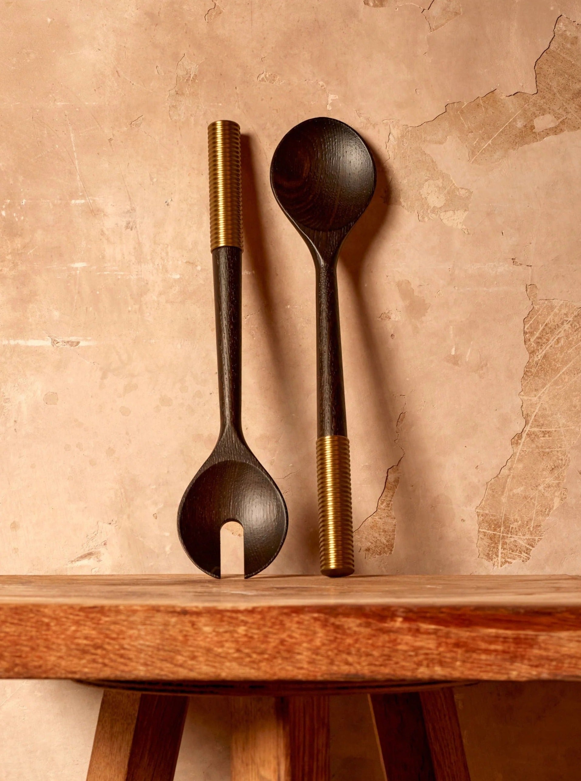 Two dark smoked ash Alhambra serving utensils with hand-carved handles rest against an aged wall, neatly positioned on a wooden shelf with visible grain. (LOBJET)